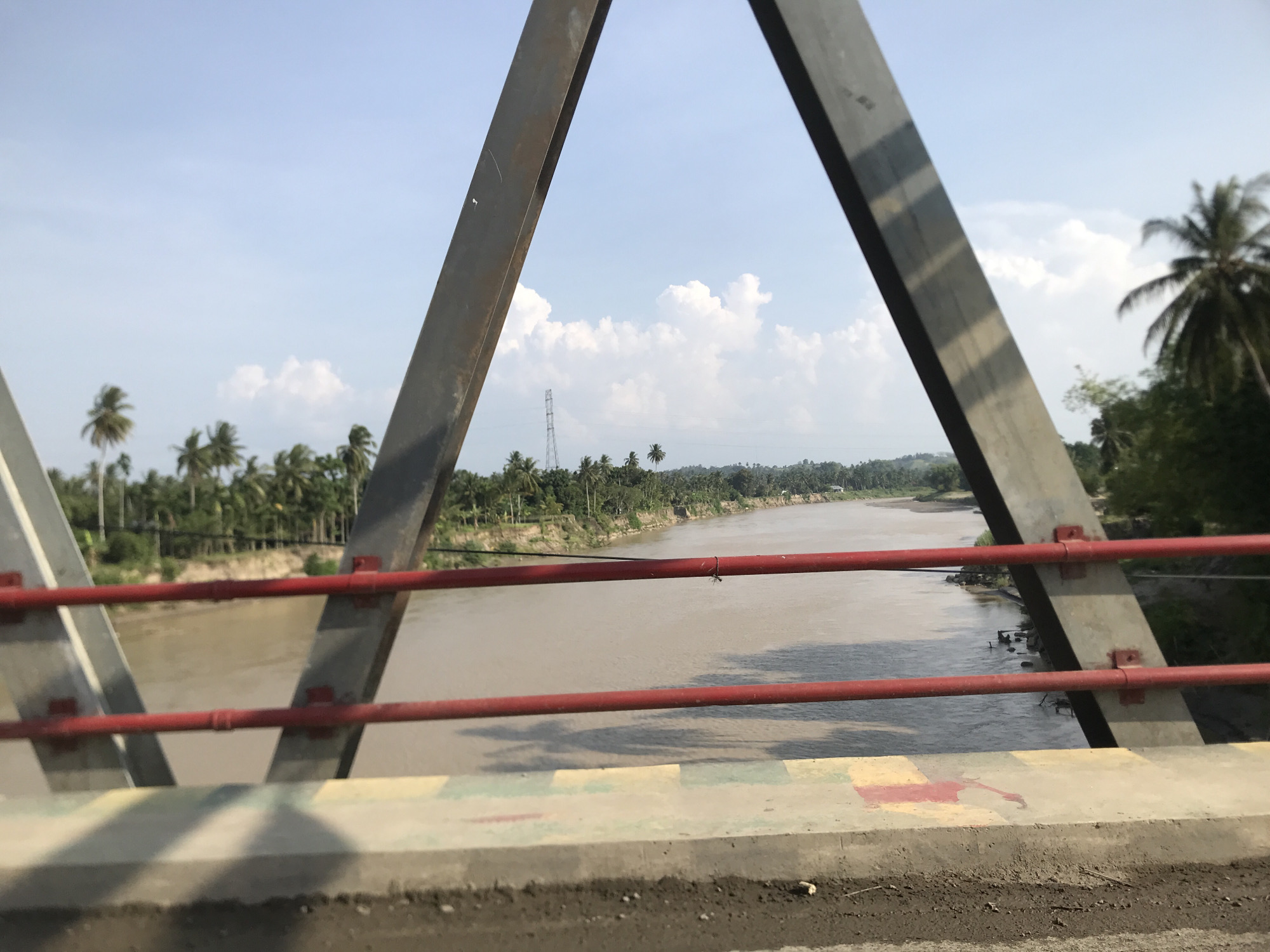 The image captures a sunlit view from a bridge crossing a wide, muddy river, set in Meuse, Indonesia. The foreground is framed by the dark gray metal truss structure of the bridge, forming a prominent "A" shape. Two red metal railings extend horizontally across the middle of the frame, serving as a safety barrier. Below these railings, a concrete barrier, adorned with faded and abstract painted designs in yellow, red, and green, forms the very bottom edge of the image.
The wide, brownish river flows from the distance towards the foreground, its surface showing reflections of the bridge structure and the sky. Both banks of the river are densely covered with lush green vegetation, primarily tall palm trees, indicative of a tropical climate. On the distant left bank, a tall metal electricity pylon or transmission tower is visible among the trees. The sky above is bright blue with scattered white cumulus clouds, suggesting clear and sunny weather during the day.
No human activity or specific event is taking place, presenting a tranquil and natural landscape. A small insect is visible on the upper red railing. The scene offers a peaceful perspective of the Indonesian countryside from the vantage point of the bridge.