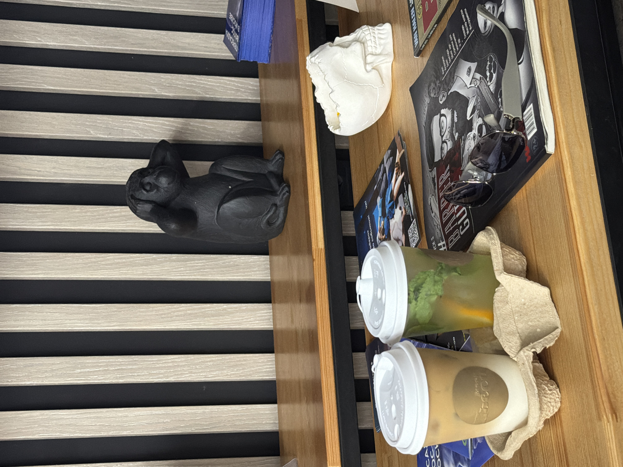 This image shows a close-up view of a wooden table with various items. In the foreground, two disposable cups with white lids are placed in a cardboard tray. The cup on the left contains a light brown beverage, likely iced coffee, with a circular logo on its side. The cup on the right holds a green beverage with mint and an orange slice.
Behind the cups, there are several magazines and a decorative white skull on the table. The skull is positioned to the right and appears to be hollow, possibly a planter or container.
Further back on the table, a black figurine of a monkey covering its ears is visible. The background features a wall with vertical wooden slats alternating with black stripes, creating a striped pattern. A stack of blue papers is also seen on the far right.
The scene appears to be indoors, possibly in a cafe or a shop, given the presence of beverages and reading materials. The lighting suggests it is daytime.
This image does not contain any information about the city of Rostov-on-Don, Russia.