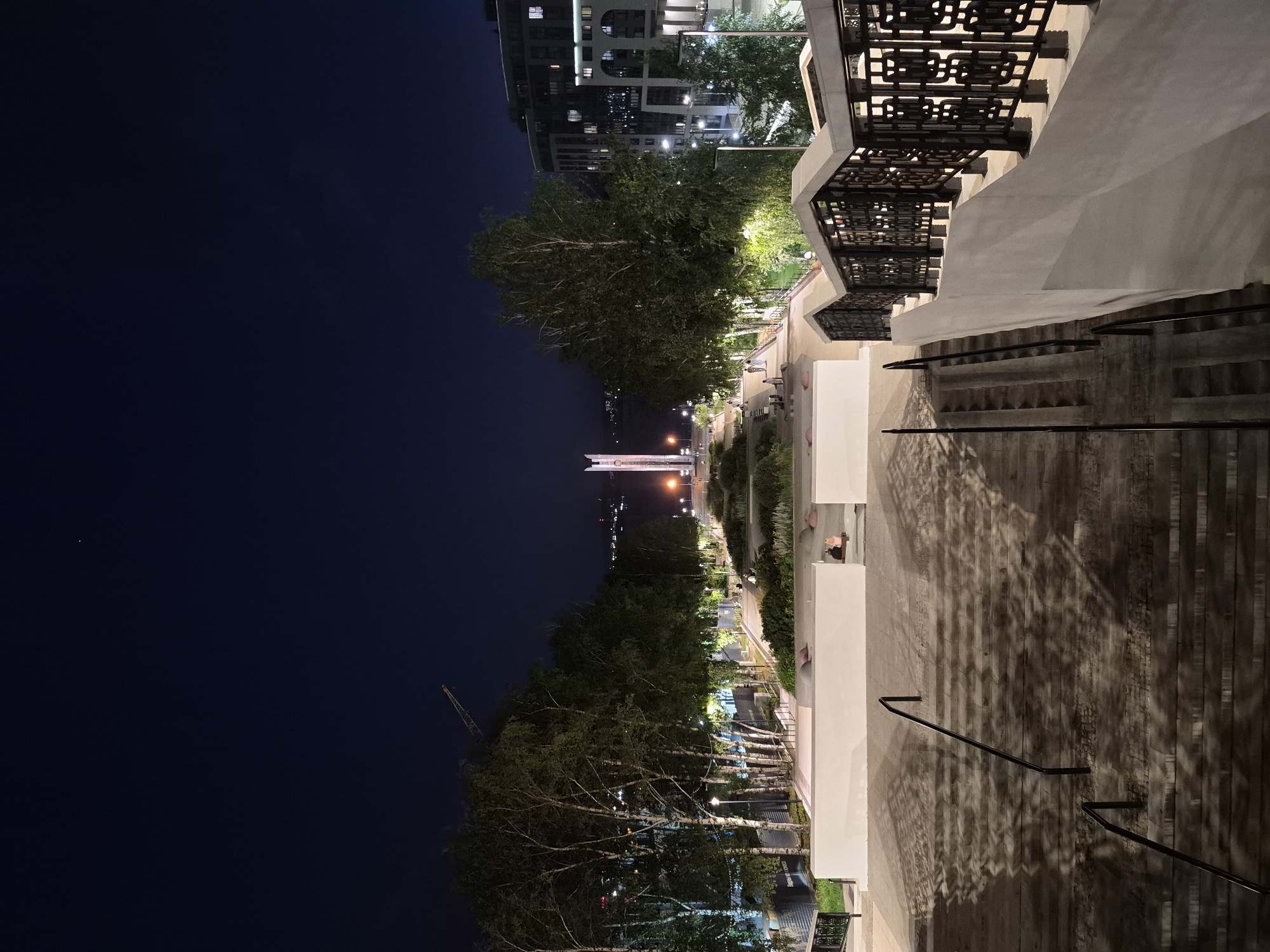 This is a nighttime, eye-level shot of a park area with a monument in the distance, located in Izhevsk, Russia. In the foreground, a wide, stone staircase descends, with black metal railings on either side. Shadows are cast across the stairs from nearby trees and decorative metalwork. To the left of the staircase, a low, white wall encloses a planted area. Further into the park, a paved walkway is lined with trees and illuminated by streetlights. Several people are visible walking or sitting in the park. Beyond the trees, a tall, slender monument stands prominently, illuminated from below. In the far distance, across a body of water, the lights of a city are visible, including the illuminated facade of a modern building on the right side of the image. A construction crane is visible to the left of the trees. The sky is dark blue and clear, with a single bright star visible near the top of the frame.