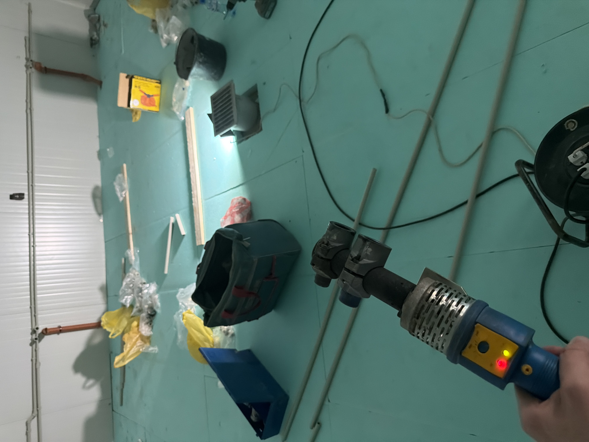 The image depicts an indoor construction or renovation site in Nikolaevo, Bulgaria. The floor is covered with pale blue insulation material. Various construction materials and tools are scattered around, including yellow plastic bags, white pipes, a blue toolbox, a green duffel bag, a black bucket, and a yellow and blue tool with red and yellow indicator lights, possibly a pipe welder. A square metal grate is visible in the floor, indicating a drain or vent. In the background, the walls are made of white corrugated panels, and pipes run along the ceiling. The scene suggests an ongoing construction project with materials laid out for use.