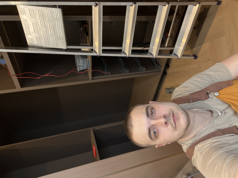 An image displays a male individual in an indoor setting, appearing to take a selfie. The man has short, dark hair and light stubble, wearing a light-colored t-shirt underneath brown and orange overalls with metal buckles. He wears a silver chain with a pendant and looks directly at the camera with a neutral expression.
The background reveals a room with partially assembled dark brown or grey cabinetry units. On a shelf within the left cabinet section, a red-handled tool is visible. In the right cabinet section, a clear container filled with fasteners or screws rests on a shelf, and below it, several metal drawer slides are installed vertically, indicating incomplete drawer assembly. A red electrical wire hangs loosely from an upper shelf. A silver-colored, four-step folding ladder with blue plastic caps on its feet and step edges is positioned on the right side of the frame. The floor is covered with light-colored wooden planks. The specific location within the city of Khimki, Russia, cannot be determined from the visual information presented.