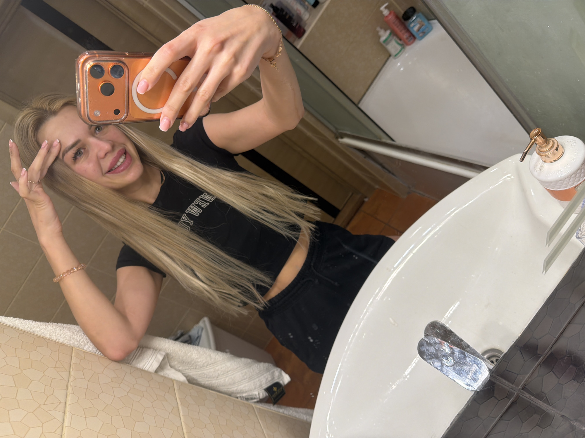 A young woman with blonde hair, wearing a black crop top and black sweatpants, takes a selfie in a bathroom mirror. She has on bracelets and rings and is smiling. The bathroom has beige tiled walls and a white sink with a chrome faucet. There are bottles of toiletries on a shelf in the background, and a white towel hangs on the wall. The scene is likely in Kyiv, Ukraine, given the context of the request.