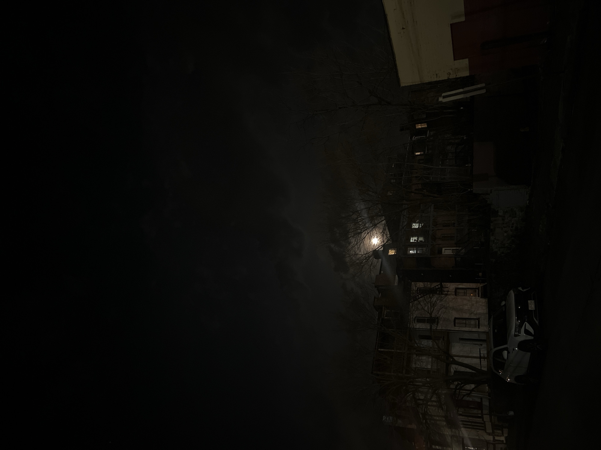 The image depicts a nighttime street scene in City of Newburgh, United States of America. Residential buildings line the right side of the frame, with windows showing faint light. A white car is parked on the street. The left side of the image is predominantly dark, with a lighter area higher up suggesting a moonlit sky partially obscured by clouds. Bare tree branches are visible in the foreground.