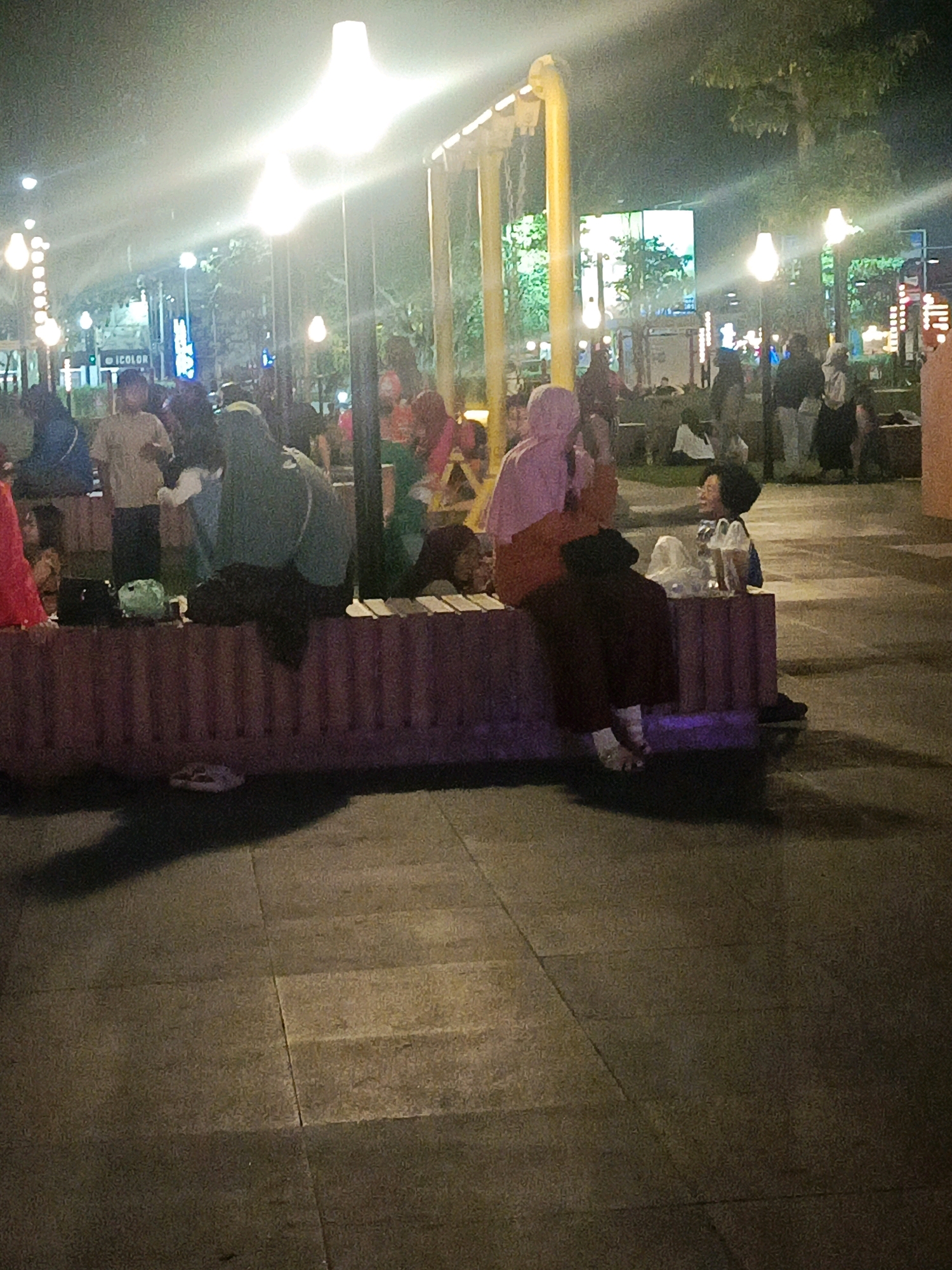 An outdoor scene at night in Sidoarjo, Indonesia, depicts a gathering of people on a paved area. Several individuals are seated on a low, segmented bench or barrier constructed of a reddish-purple material. Some are seen from the side, while others are facing the viewer. A person in a light-colored hijab and orange clothing is seated to the right, facing a person with dark, curly hair wearing a blue garment. In the background, a large, bright screen is visible, along with various illuminated structures and trees. Several tall lampposts with bright lights illuminate the area, casting shadows on the ground. A child in a light-colored shirt is standing and looking towards the camera on the left side of the frame, while other figures are seated or standing further back. The ground is paved with large, rectangular tiles.