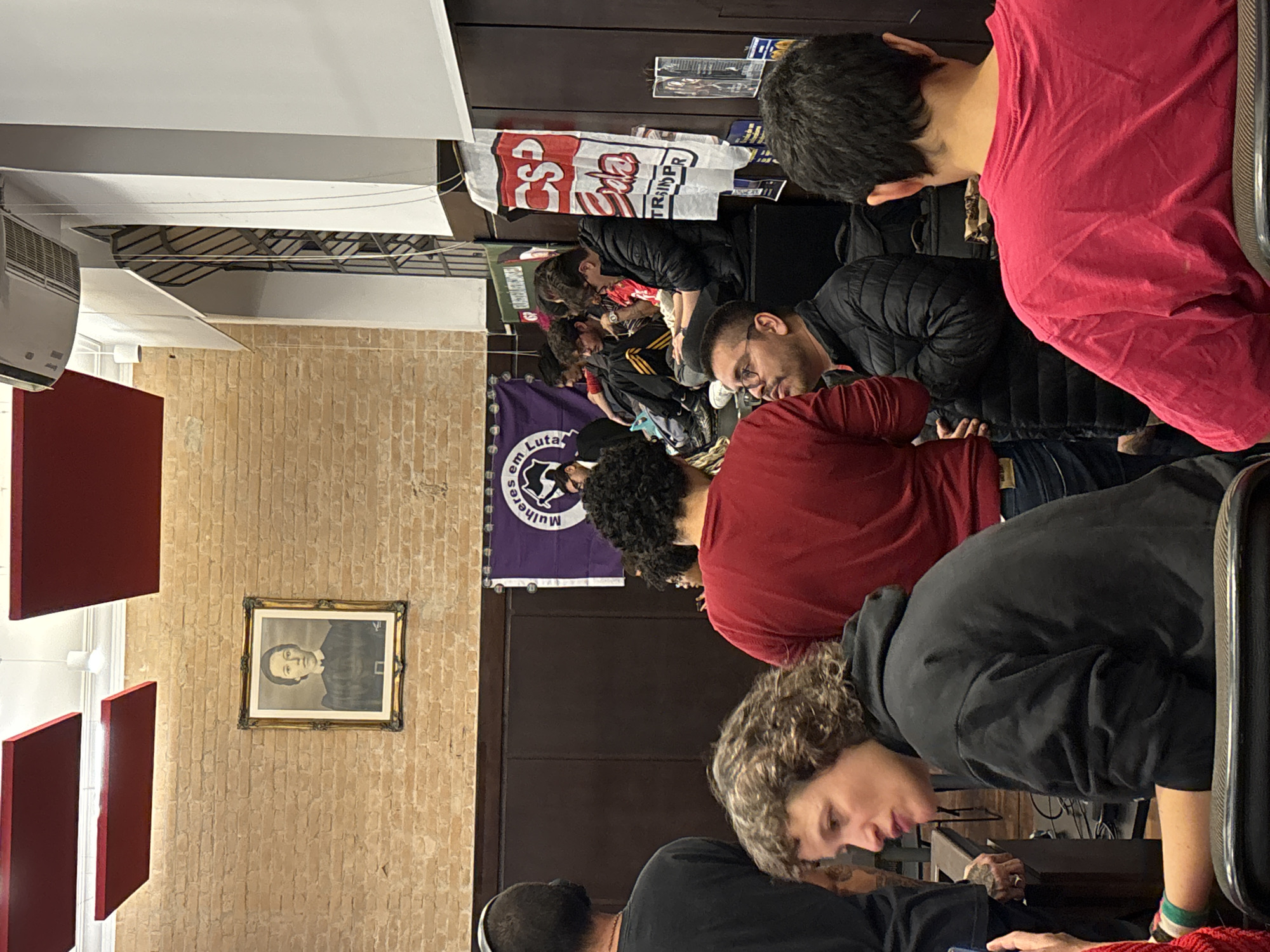 A medium shot shows a group of people gathered indoors in what appears to be a meeting room. The room features a brick wall adorned with a framed black-and-white portrait and red acoustic panels. Banners and flags are visible in the background, including one with the text "CSP-Conlutas TRINCHEIRA P..." and another purple banner with text in Portuguese, "Mulheres em Luta." The room appears to be located in the city of São Paulo, Brazil.
The individuals are diverse in appearance, some with dark hair and others with curly or wavy hair. They are dressed in casual clothing, including red shirts, black jackets, and hoodies. Most are looking down, possibly engaged with devices or taking notes. The scene suggests a discussion or presentation in progress.