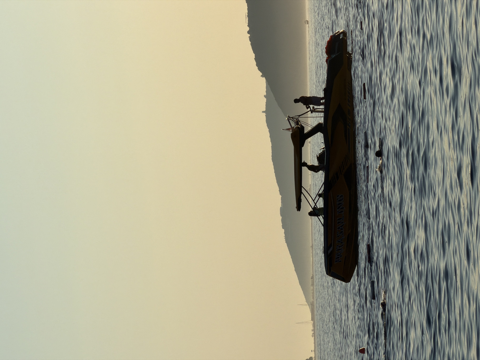 A silhouette of a yellow and black speedboat with "PARASAIL LINES" written on its side floats on choppy blue water. Three figures are visible on the boat, two appear to be standing, and one is seated. In the background, a hilly coastline is visible under a pale yellow and blue gradient sky. Several buoys and floating objects are scattered in the water in front of the boat. The scene is likely a recreational area, possibly a beach or bay, in Boreti, Montenegro.