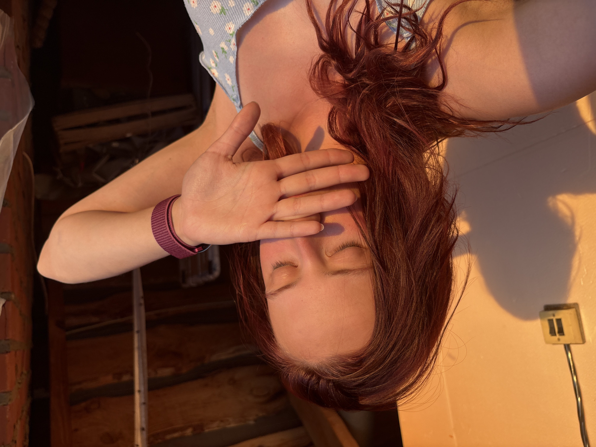 A woman with auburn hair covers her face with her right hand. She is wearing a light blue, floral-patterned top. A purple fitness tracker is on her left wrist. Behind her to the left is a brick wall and a wooden staircase. To the right, a white wall with a light switch is partially illuminated by an orange glow. The location is presumed to be indoors. The image appears to be taken in Прикамский, Russia.