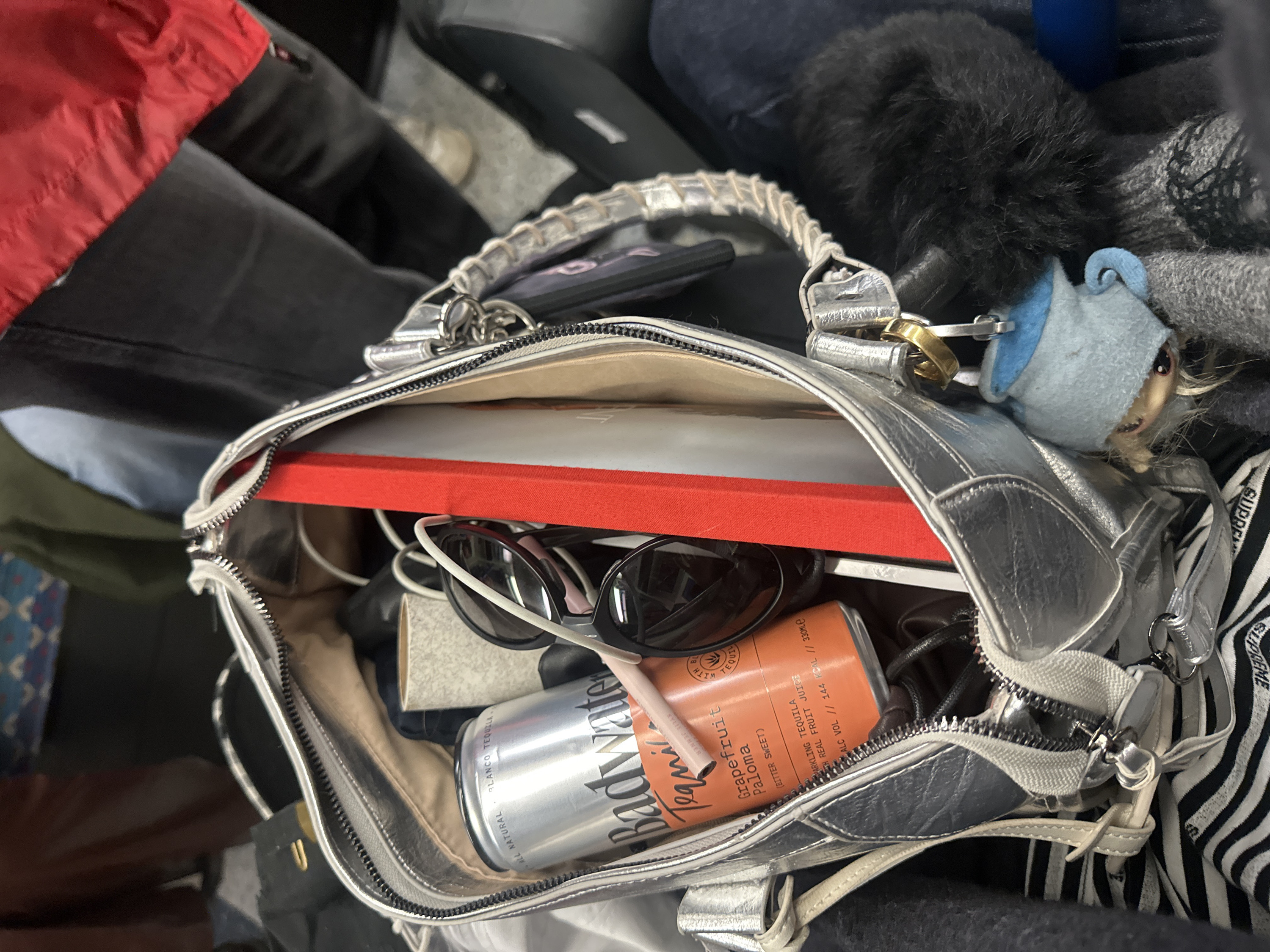 The image provides an overhead view into an open silver metallic handbag, revealing its contents packed together. The bag itself has braided or woven handles and a zippered opening.
Inside the bag, several items are visible:
* A large, flat, rectangular object with a vibrant red cover or spine is placed horizontally across the bag. This could be a book, a tablet, or a planner.
* A pair of black sunglasses with thick white or light-colored frames is resting on top of other items.
* Two beverage cans are prominently featured. One is silver with black text, partially visible, reading "ALL NATURAL", "BLANCO TEQUILA", and part of a brand name, which appears to be "Body water". The second can, colored orange, clearly displays "Body water Tequila", "Grapefruit Paloma", "(BITTER SWEET)", "SPARKLING TEQUILA", "WITH REAL FRUIT JUICE", and "ALC VOL // 144 KCAL // 330MLE".
* Next to the cans, there is a small, beige or light brown rectangular object, possibly a small notebook or wallet.
* A white pen-like object with "KYLIE" faintly visible on it is seen near the orange can, likely a cosmetic item.
* Attached to one of the bag's zipper pulls or straps is a small, light blue plush doll or keychain with blonde hair and large eyes, wearing a pointed hat.
* A black, fluffy or furry object is visible near the top right edge of the bag, possibly a keychain or part of a garment.
* A black smartphone with a purple-patterned case is partially tucked into the left side of the bag.
* Metal keyrings and a gold-colored ring are attached to the bag's handle or zipper.
* The bag's interior lining appears to be a light, neutral color, and a black and white striped fabric with the word "SUPREME" is visible towards the bottom right.
The background around the bag is blurred and consists of dark, indistinct shapes, suggesting other luggage, clothing, or a car seat. A vibrant red piece of fabric, possibly a jacket or garment, is visible in the upper left background.
The overall scene suggests a personal bag packed for travel or a busy day, containing essentials for leisure and convenience.