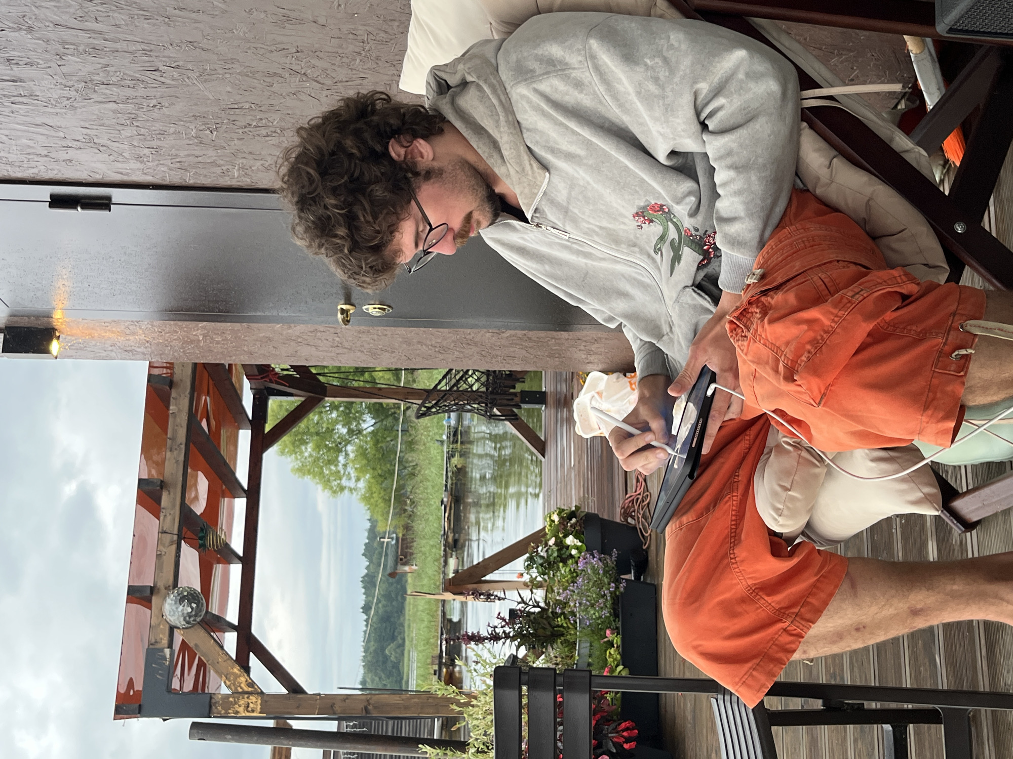 In Radchenko, Russia, a young man with curly brown hair and glasses is seated in a chair on a wooden deck. He is wearing an orange pair of shorts and a grey hooded jacket with a green serpent and floral design. He is holding a tablet in his hands and appears to be drawing on it with a stylus. The background shows a calm body of water surrounded by lush green vegetation and trees, with a cloudy sky overhead. A wooden structure with a red translucent roof is visible to the left, adorned with a disco ball and a small hanging object. The scene conveys a sense of relaxation and creative engagement in an outdoor, possibly lakeside, setting.