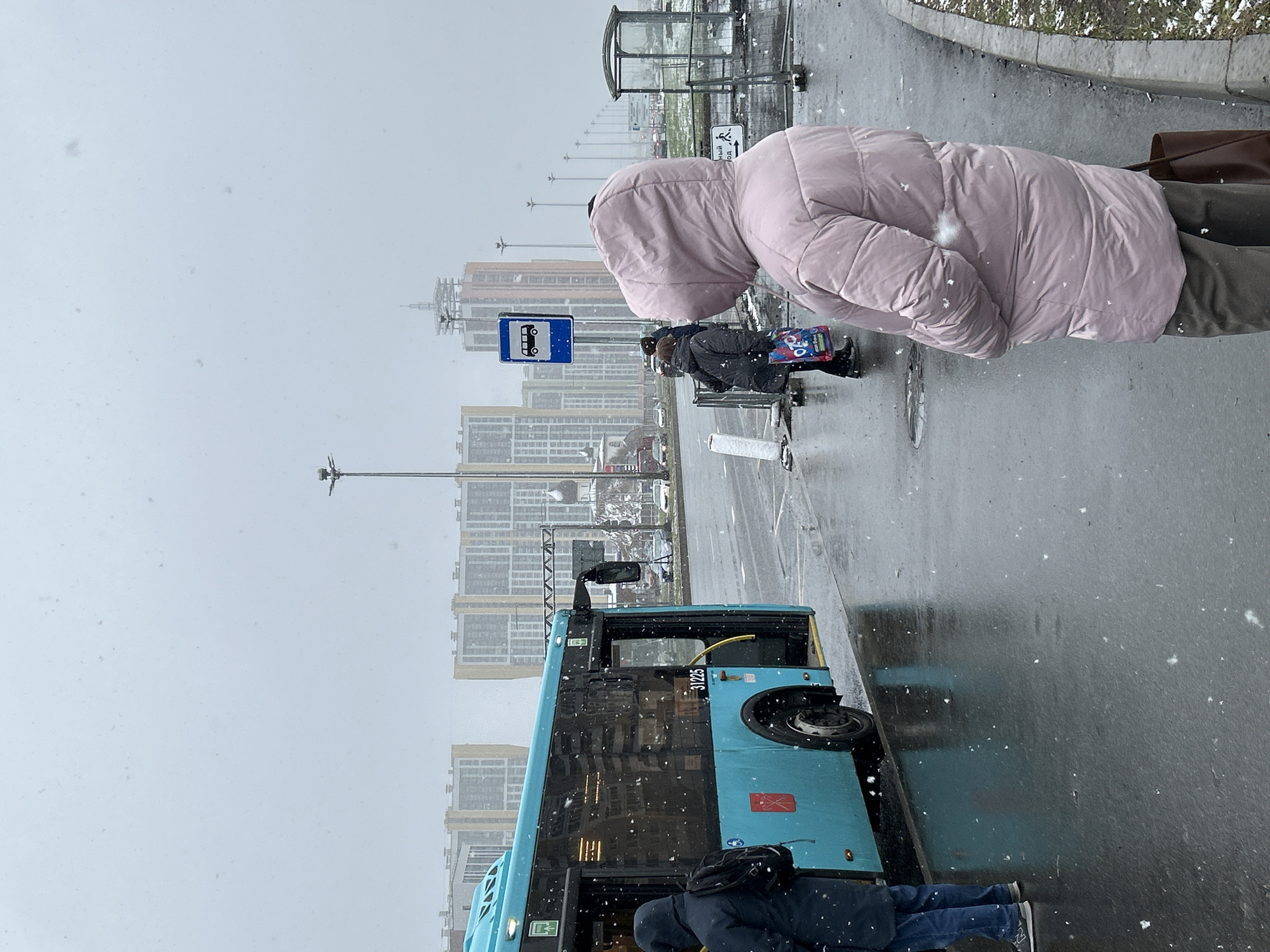 The image captures a snowy day at a bus stop in what appears to be Saint Petersburg, Russia. A blue bus is visible, partially obscuring the view of a modern residential area in the background. Several people are present. One person in a pink hooded jacket stands prominently in the foreground. Other individuals are standing at the bus stop, possibly waiting for the bus. The ground is wet, likely from melting snow. A bus stop sign is visible along with other urban infrastructure elements, like lampposts and metal bus shelters. The sky is overcast, suggesting ongoing snowfall.