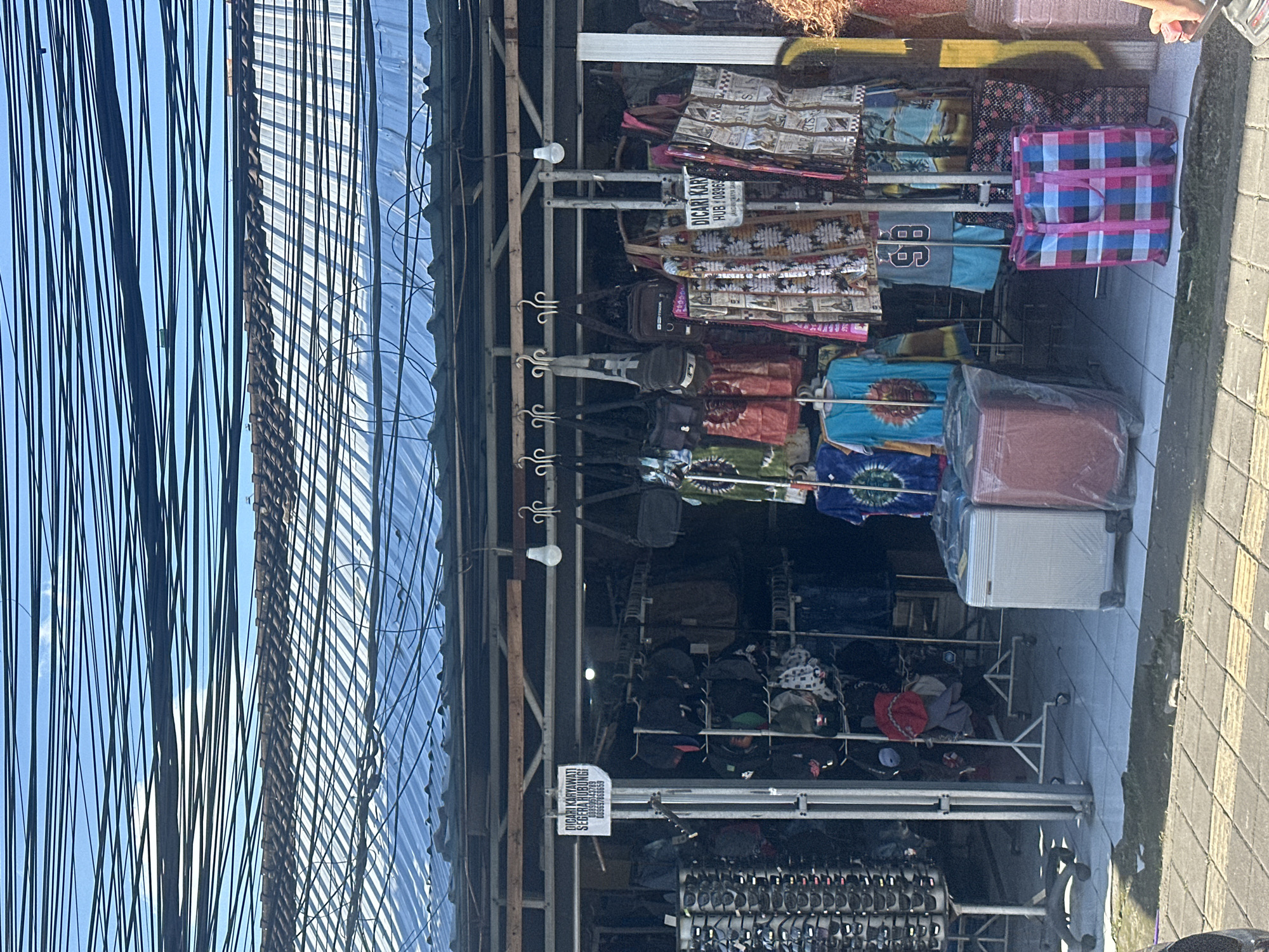 The image depicts the storefront of a small shop in North Kuta, Indonesia. The shop appears to sell clothing, bags, and possibly sunglasses, with items displayed on racks and hanging from the ceiling. A sign in Indonesian, which translates to "Looking for employees! Contact immediately!", is prominently displayed. In front of the shop, two suitcases are visible, one white and one red, both covered in plastic. A colorful checkered tote bag is placed on the sidewalk. The environment is characterized by an overhead canopy of wires and a tiled floor. The sun casts shadows on the ground, indicating it is daytime. There are no people clearly visible interacting within the scene.