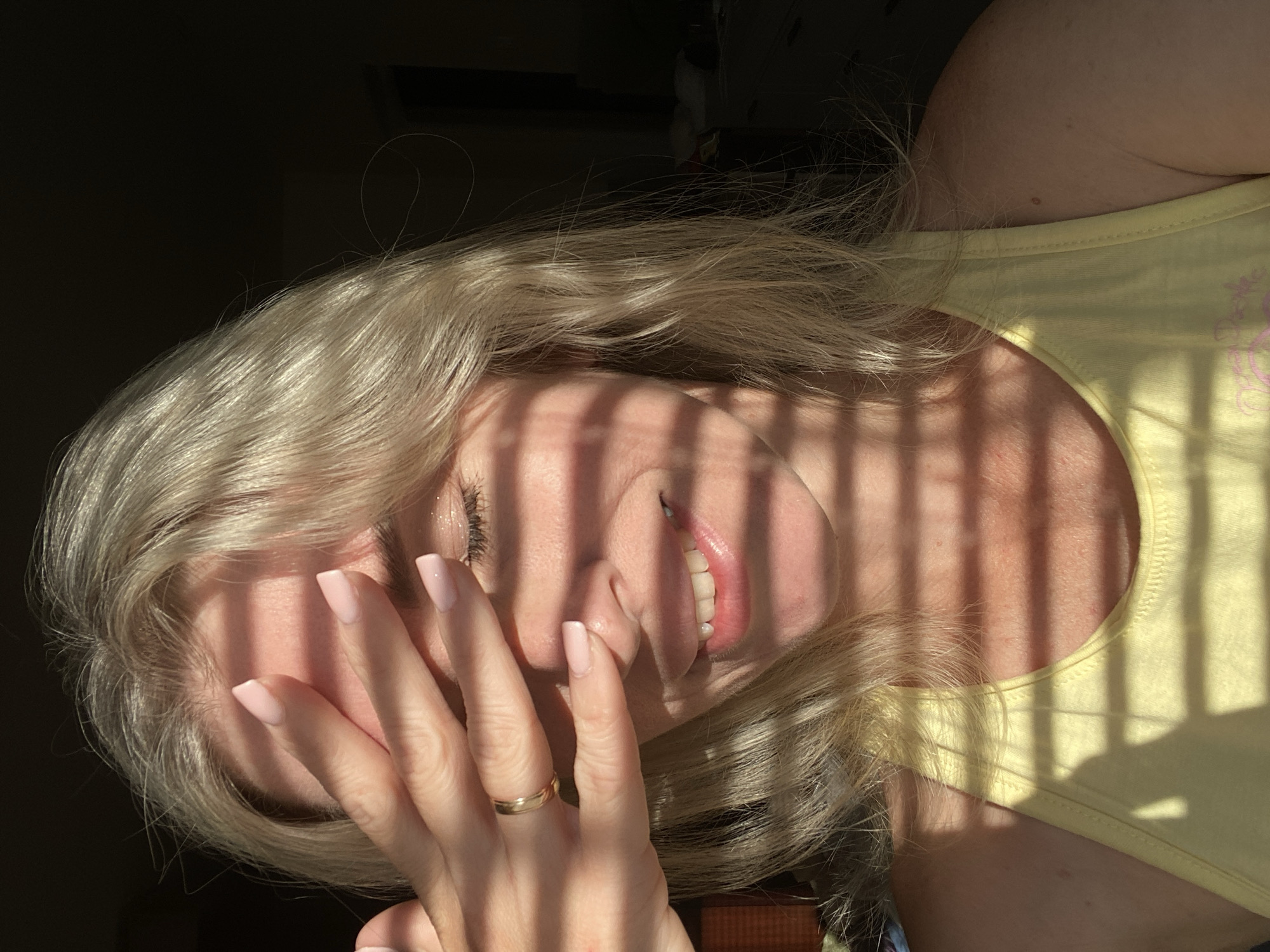 In an indoor setting, possibly a bedroom, a person with blonde hair, partially obscured by their hand, is captured in a close-up portrait. Sunlight streams through blinds, casting horizontal shadows across their face and upper body. The individual is wearing a yellow tank top and a gold ring is visible on their hand. The background is dark, with hints of furniture and a dresser. The overall lighting suggests it is daytime. The location is Unknown, Unknown.