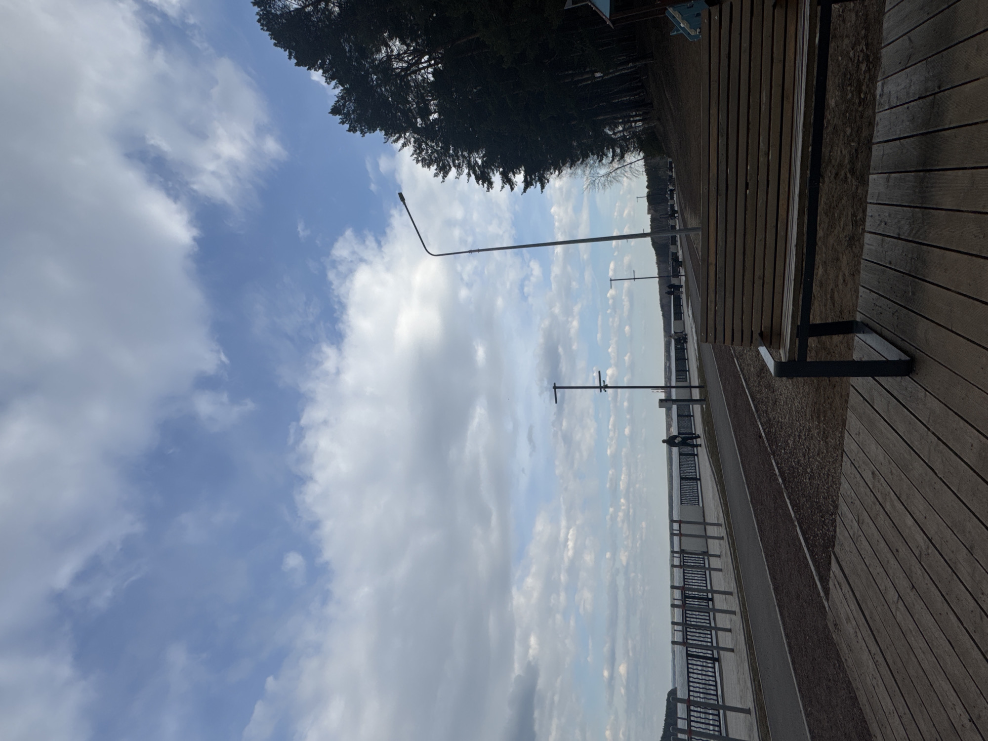 The image captures a scenic waterfront promenade in Chaikovsky, Russia, on a partly cloudy day. The sky dominates the upper portion of the frame, filled with a mix of fluffy white and grey clouds against patches of clear blue. Along the horizon line, a dense forest of dark evergreen trees runs, suggesting a natural boundary to the area.
In the middle ground, a wide body of water, likely the Kama River reservoir, stretches into the distance under the sky. A paved path or sidewalk runs parallel to the water, separated by a black metal railing. Several tall lampposts are spaced along this path. A few small figures of people can be seen walking in the distance, enjoying the promenade.
In the foreground, a wooden boardwalk with visible planks occupies the right side of the image, leading towards the water. A sturdy wooden bench with black metal supports is prominently placed on this boardwalk, inviting relaxation. To the left of the boardwalk and parallel to the paved path, there is a strip of gravel or dirt.
The scene conveys a peaceful and expansive outdoor setting. It is daytime, with ample natural light indicating fair weather, despite the clouds. No discernible text is visible in the image.