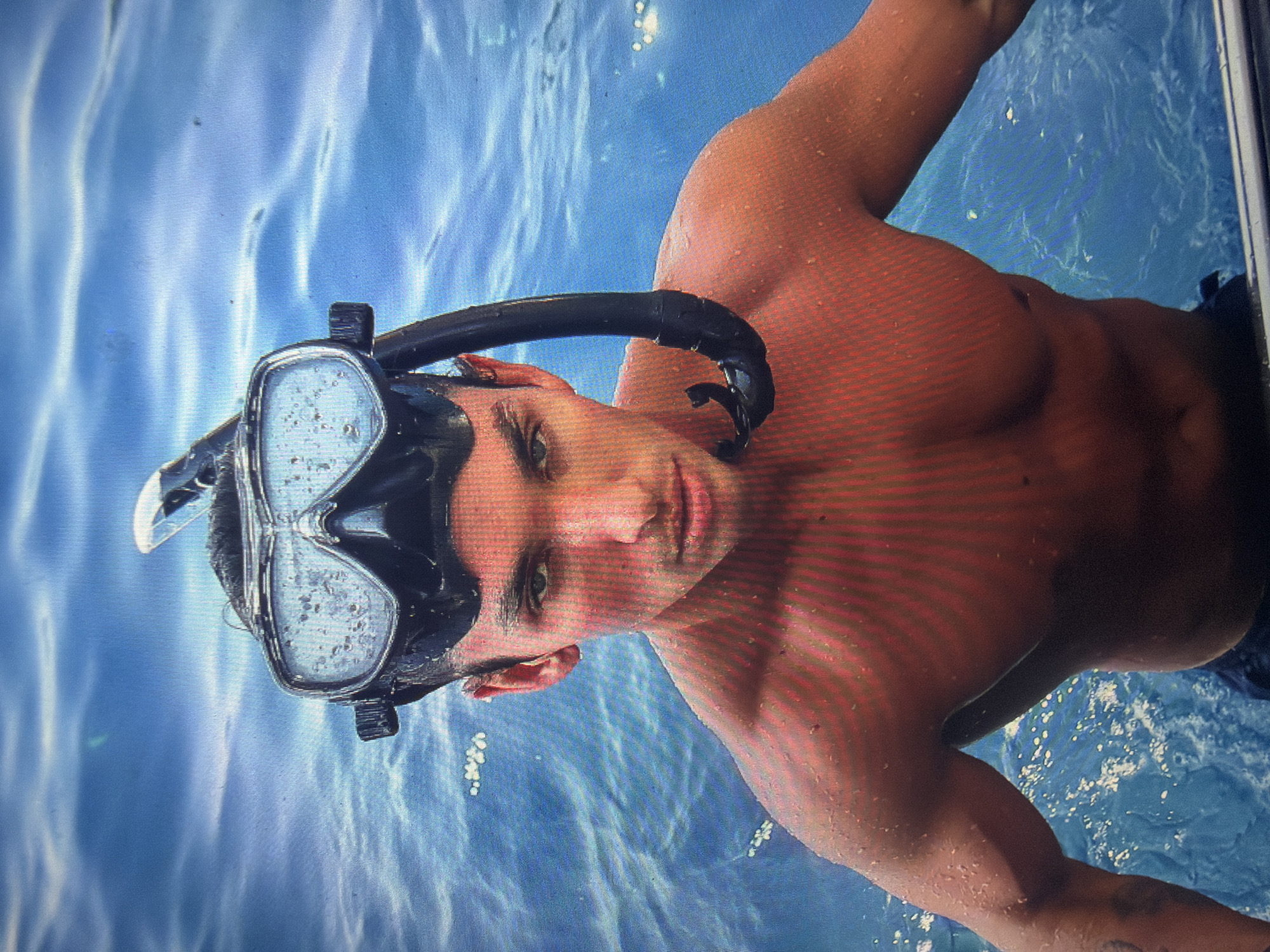 The image depicts a young adult male partially submerged in clear blue water, a scene situated in the city of Shumen, Bulgaria. The subject, with fair skin and dark hair, directly engages the viewer with his gaze. He wears a black snorkeling mask positioned on his forehead, revealing his eyes, and a black snorkel extends along his right shoulder. Numerous water droplets cling to his skin and hair, indicating recent submersion. His bare upper body, including chest and shoulders, exhibits a toned physique with visible musculature. The background is entirely composed of vibrant blue water, characterized by surface ripples and reflected light, suggesting an outdoor, sunlit environment. A light-colored edge or rail is partially visible at the bottom right corner of the frame.