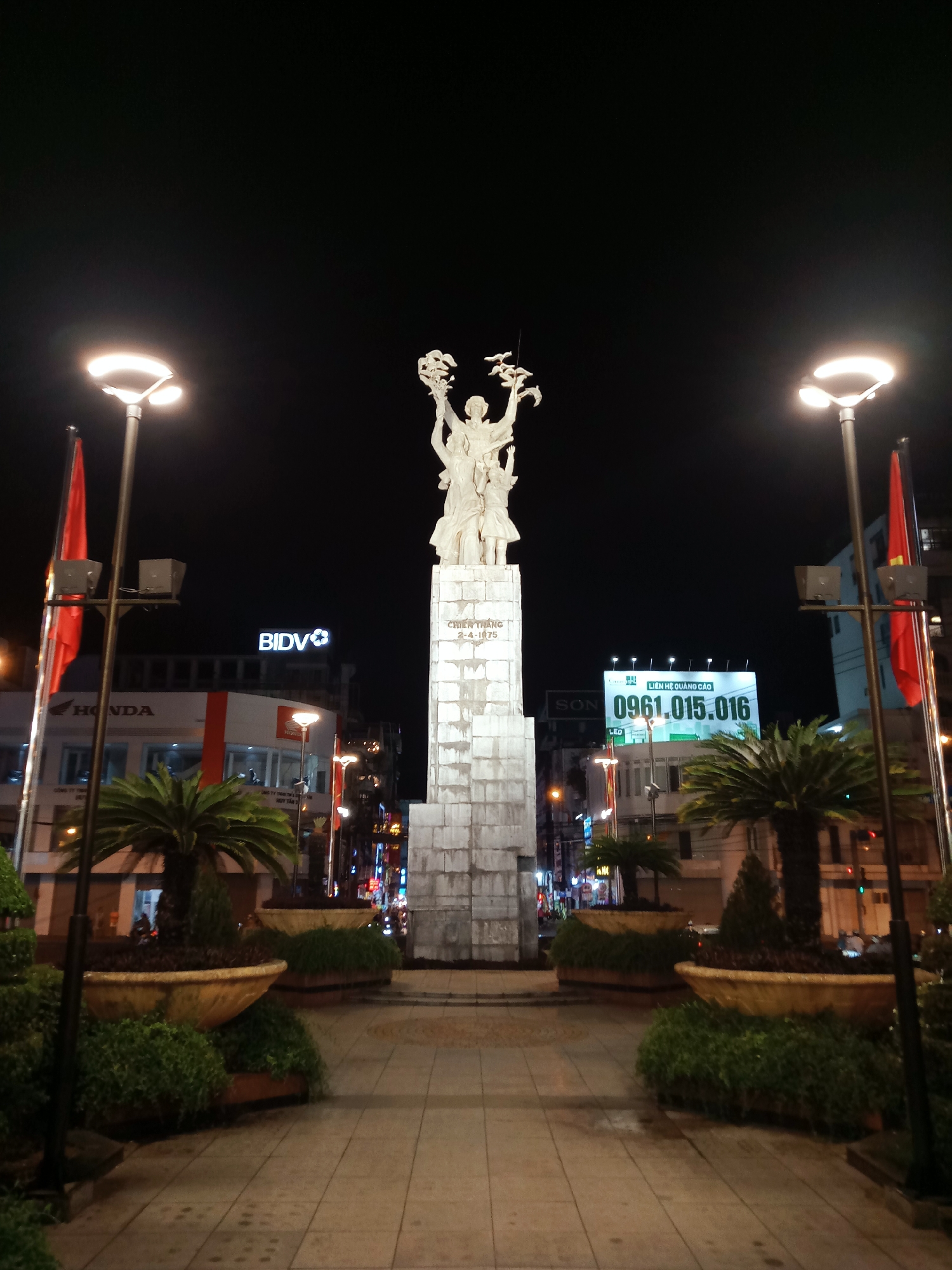 The image presents a nighttime view of a public square or memorial area in Nha Trang Ward, Vietnam. A prominent, tall white stone monument stands centrally, topped by a statue depicting several figures, possibly a family, holding what appears to be a wreath or a plant with doves aloft. The monument's base is inscribed with "CHIẾN THẮNG 2-4-1975," which translates to "VICTORY 2-4-1975."
Two modern streetlights illuminate the scene from the sides, and red Vietnamese flags are displayed on poles next to them. The foreground features a paved walkway, flanked by large decorative planters filled with green shrubs and tall palm trees, creating a landscaped pedestrian zone.
In the background, behind the monument, various commercial buildings are visible, lit by their own internal and external lights. Visible signs include "HONDA" and "BIDV" (a Vietnamese bank) on the left side. On the right, a large billboard displays Vietnamese text "LIÊN HỆ QUẢNG CÁO" (Contact for advertising) and a phone number "0961.015.016," along with smaller text "SƠN" and "LED." The clear night sky and the artificial lighting contribute to a serene urban atmosphere. No specific activity or event is taking place, making it a static depiction of a city landmark at night.
