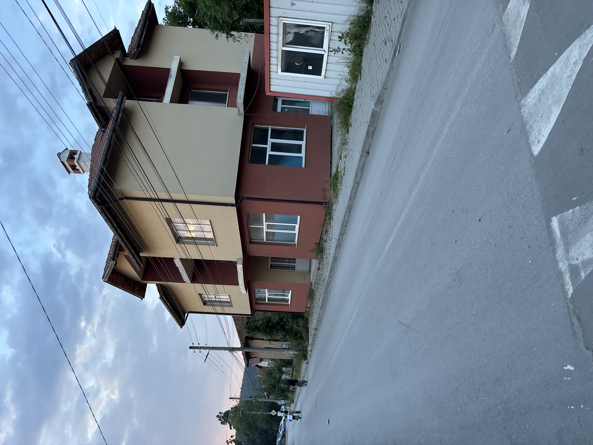 The image depicts a residential street scene at dusk or dawn in an unknown location. A two-story building dominates the right side, featuring a cream-colored upper facade and a reddish-brown lower facade, a tiled gabled roof with a chimney, and multiple windows. A small white, possibly corrugated metal, structure with a window stands adjacent to the main building on the right. The asphalt road in the foreground has a white dashed center line and a triangular white road marking, likely part of a crosswalk. A concrete sidewalk lines the right side of the street, with sparse vegetation growing through cracks. On the left side of the street, utility poles with numerous power lines are visible, extending above the main building. Further down the street, older, smaller buildings are obscured by trees and foliage. Two individuals on bicycles are riding away from the camera on the left side of the road; one is on a smaller bicycle or scooter, and the other on a larger bicycle. A yellow diamond-shaped road sign is mounted on a pole further down the street. In the distant background, a mountain range is visible under a sky with scattered clouds.