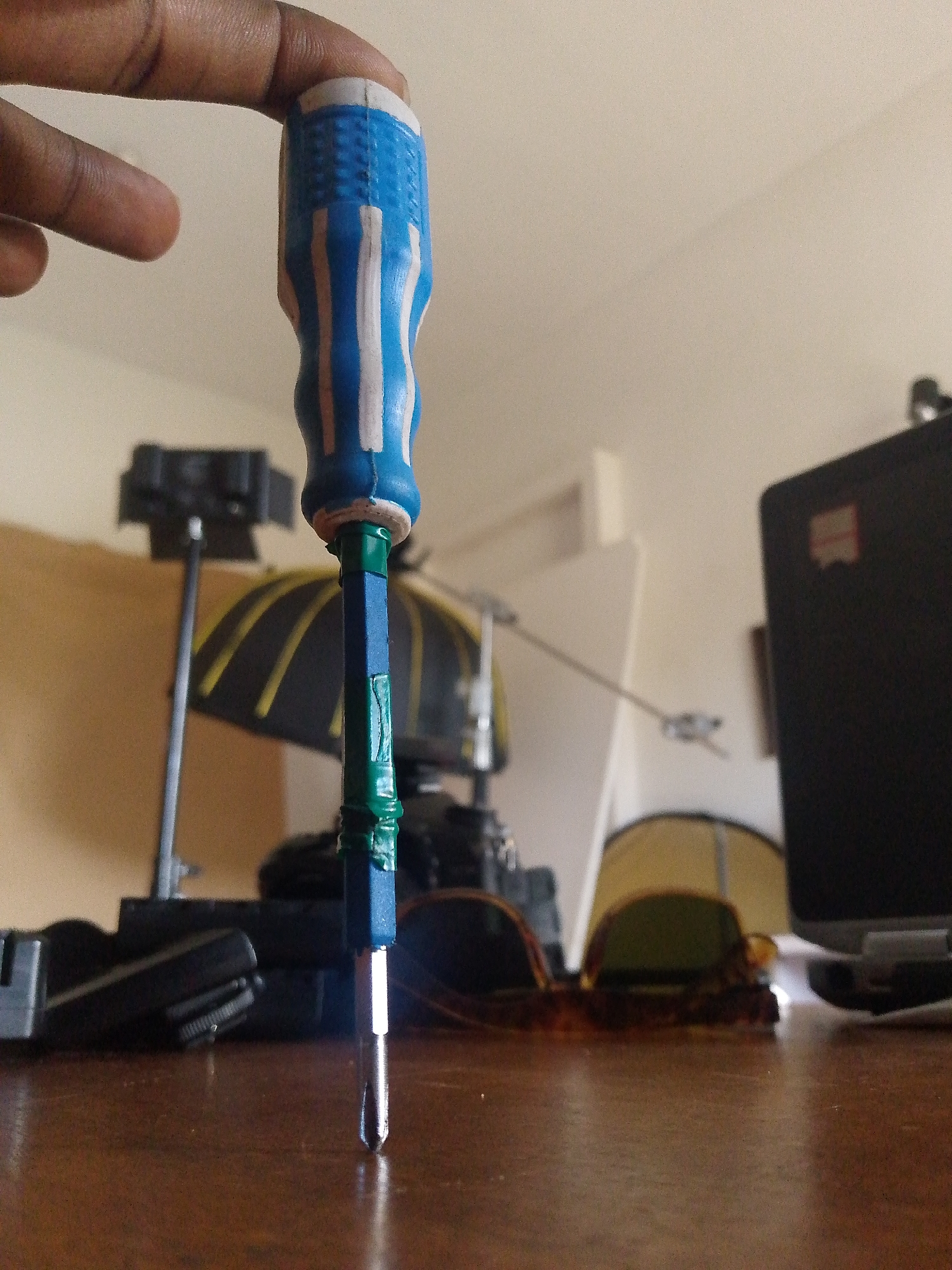 A close-up, low-angle shot shows a person's fingers holding the handle of a screwdriver. The screwdriver has a blue and white striped handle. The shaft of the screwdriver is blue and wrapped with green tape. The tip of the screwdriver is silver and pointed. The screwdriver is held vertically, pointing downwards towards a wooden surface.
In the background, the scene is blurred. To the left, there is a dark object with a handle, possibly a camera or some other equipment. Behind it, a large object with yellow and black stripes is visible, potentially an umbrella or a reflector. Further in the background, there is a light-colored wall and a white object that could be a screen or a partition. To the right of the screwdriver, a black laptop is partially visible, with a small red logo on its lid. In the middle ground, a pair of sunglasses with a tortoiseshell pattern rests on the wooden surface. A yellow tent-like structure is visible behind the sunglasses, and a thin wire extends across the background.
The lighting suggests an indoor setting, possibly a studio or a room with artificial lights. There is no indication of the time of day or weather conditions. The focus is on the screwdriver and the hand holding it, creating a sense of detail and directness. No text is clearly visible in the image.