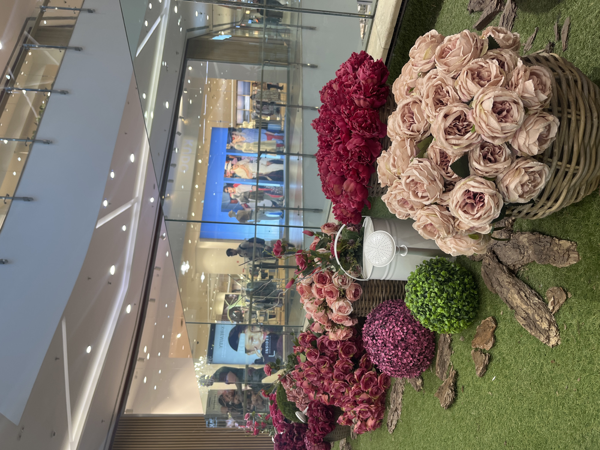 The image captures a decorative display featuring artificial flowers and greenery set against the backdrop of a modern shopping mall interior. The display includes arrangements of pink and red flowers in woven baskets, a white watering can, a purple spherical bush, and a green spherical bush, all placed on artificial green turf. Pieces of bark are scattered around the display.
The backdrop shows a brightly lit shopping mall with glass barriers, escalators, and retail displays. Banners and screens displaying advertisements are visible. The reflection in the glass suggests the presence of shoppers and other mall activities.
Based on the visual style of the retail environment and the Cyrillic alphabet possibly visible on the advertising screens, it is plausible that this scene is located in a shopping mall in Sofia, Bulgaria. However, without specific signage or location markers, the exact location cannot be definitively determined.