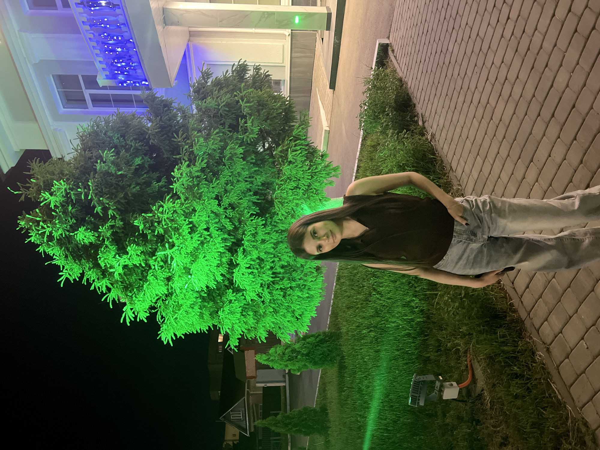 A woman stands on a brick walkway next to a strip of grass. Behind her is a building with a blue-lit overhang. A large, green-lit tree is also visible. Other buildings and trees are visible in the distance. The image was taken at night in Mendeleyevsk, Russia.