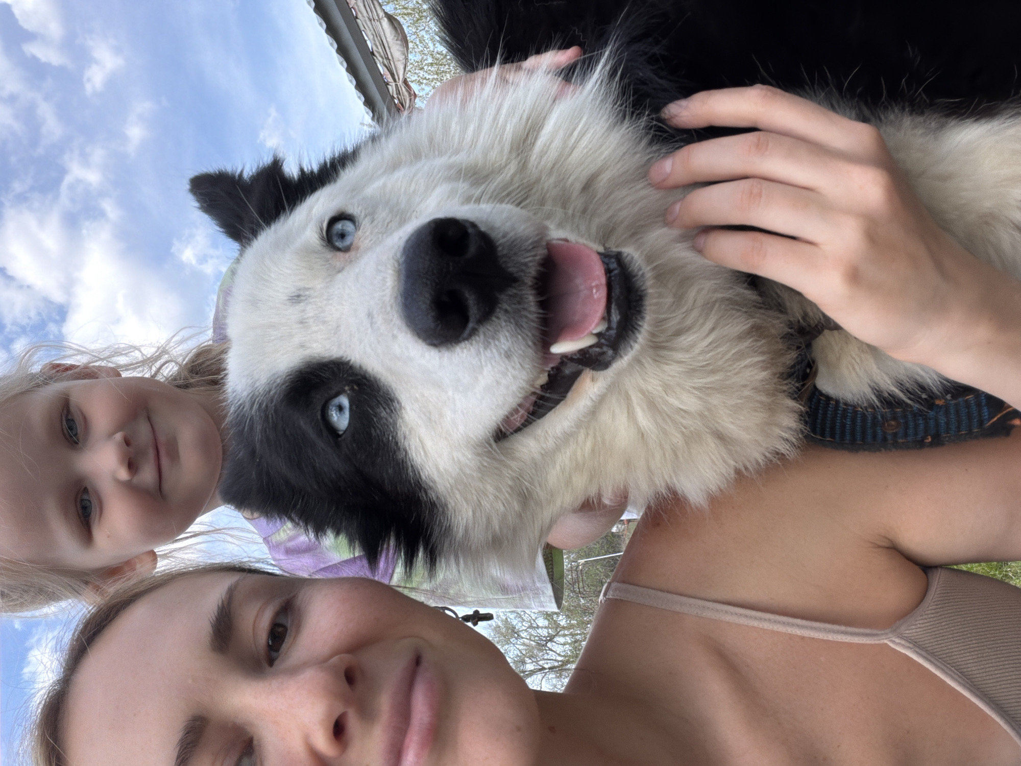 A low-angle selfie depicts an adult woman and a young girl, likely a mother and daughter, posing with a medium-sized, black and white dog with blue eyes. The woman is in the foreground with a neutral expression. The girl is positioned behind the dog and to the left of the woman. The dog is the central focus, held by a hand. The background shows a partly cloudy sky and a section of a roof. The location is possibly a residential area. There is no indication of the city Kamyshin, Russia, in the image.
