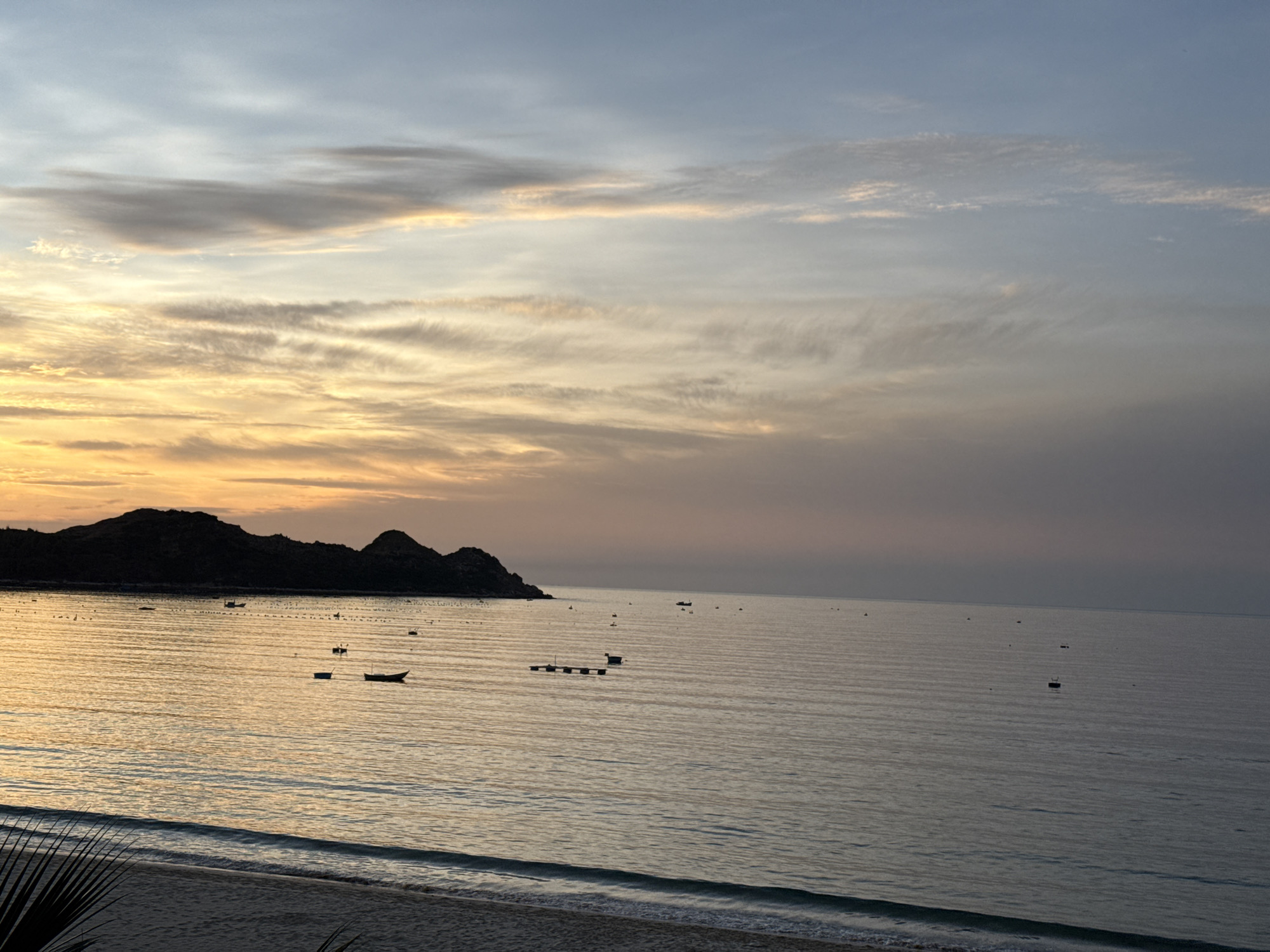 The image depicts a serene sunset over the ocean in Song Cau, Vietnam. The sky is painted with hues of orange and yellow near the horizon, transitioning into soft blues and grays higher up, with wispy clouds stretching across. A dark, silhouetted mountain range forms the backdrop to the left, with the calmer waters of the sea in the foreground. Several small boats are scattered across the water, some appearing to be fishing vessels. A sandy beach with gentle waves lapping at the shore is visible at the bottom of the frame, with a hint of palm fronds in the lower left corner. The overall atmosphere is one of tranquility and natural beauty.
