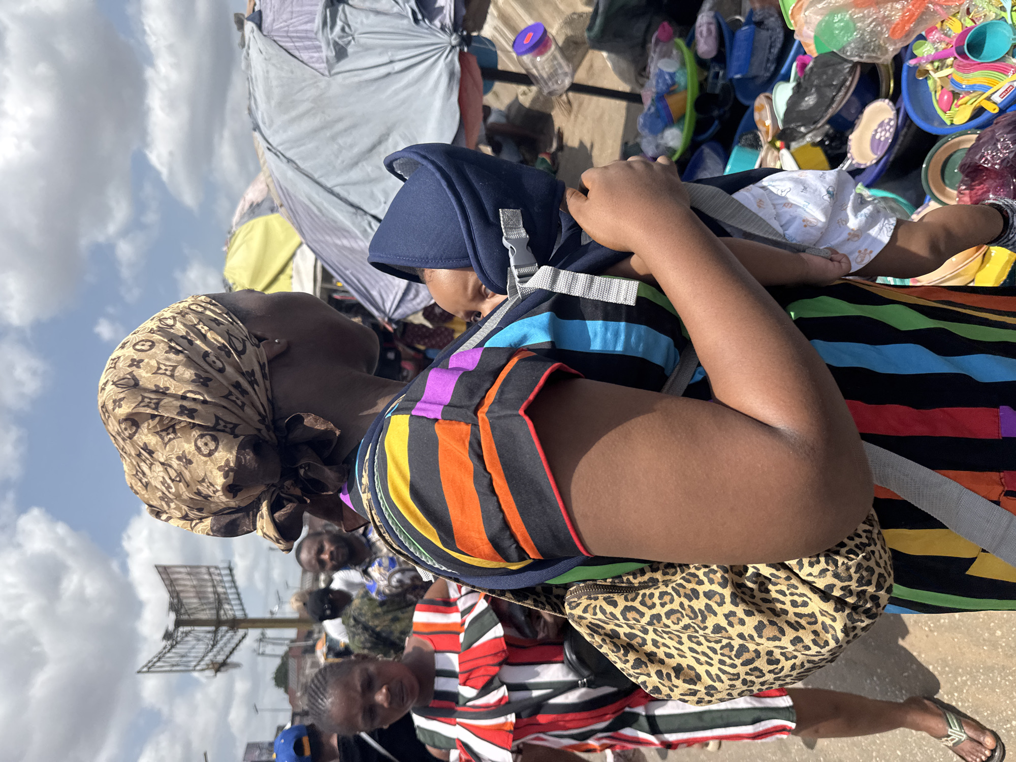 This vibrant, sunlit photograph captures a bustling outdoor market scene in Onitsha, Nigeria, under a partly cloudy blue sky. The main subject is a woman with dark skin, seen carrying a baby in a front carrier. She wears a light brown headscarf adorned with a repeating monogram pattern, a striking multi-colored striped shirt featuring black, orange, blue, purple, and green stripes, and flip-flops. A leopard print bag is slung over her shoulder. The baby, nestled in a blue carrier, is dressed in a white outfit with subtle patterns.
The background reveals the lively activity of the market, with various colorful plastic goods such as cups, plates, and utensils displayed for sale, likely on ground mats or within containers. Grey tarpaulins or umbrellas indicate market stalls providing shade. Other individuals are partially visible in the crowded background, including a young girl with braided hair in the lower foreground. The overall setting suggests a busy commercial hub within a dense urban environment. The time of day is clearly daylight with bright sunshine. No discernible text is visible other than the distinct pattern on the woman's headscarf.
