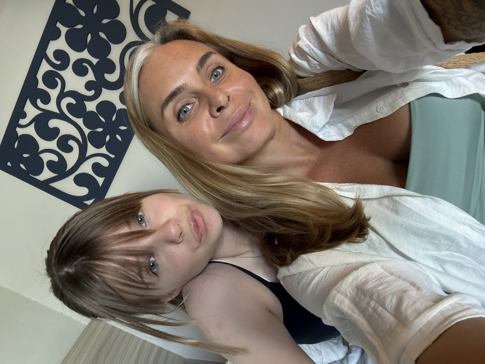 A selfie taken by a woman in Racha Thewa Subdistrict, Thailand, shows her and a young girl. The woman, who is in the foreground, has blonde hair and is smiling. She is wearing a white shirt over a light green top. The girl is in the background, leaning towards the woman. She has blonde hair with bangs and is looking towards the camera with her lips pursed. A decorative panel with a blue floral pattern is visible on the wall behind them. The setting appears to be a bedroom.