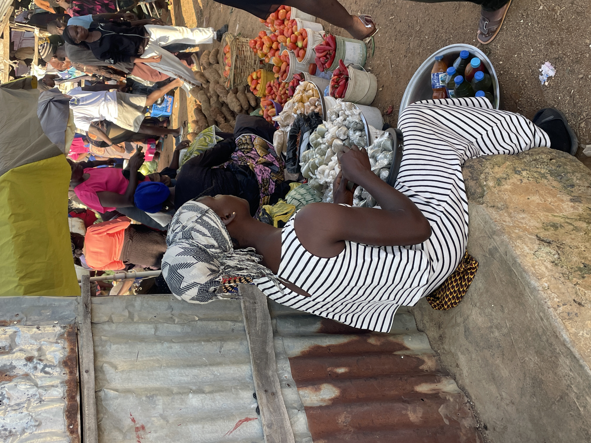 In Gura Top, Nigeria, a woman with a patterned headscarf and a black and white striped dress sits on a concrete ledge, facing away from the camera. She is surrounded by various produce and goods for sale at an outdoor market. In front of her are several bowls filled with small, green-wrapped items. To her right, a large metal basin holds several plastic bottles of liquid, some with blue caps. In the background, other market-goers are visible, along with stalls displaying an assortment of items including tomatoes, chili peppers, and what appear to be yams. The market appears bustling with activity. The overall environment is dusty and arid, with corrugated metal structures and tarpaulin coverings providing shade.