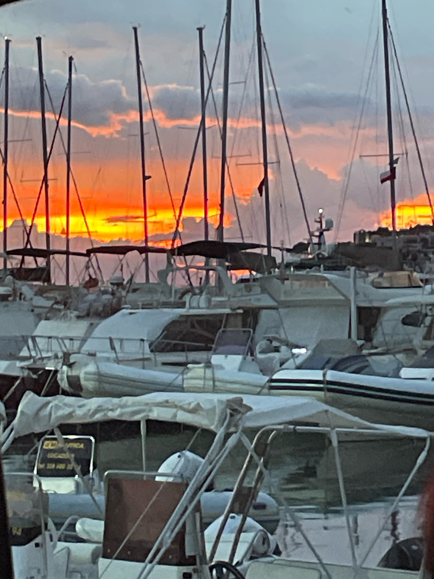 The image captures a marina at sunset, with the sky ablaze with vibrant orange and yellow hues, reflecting on the water and silhouetting the masts of numerous sailboats. The boats are docked in rows, some covered with white tarpaulins. The scene appears to be in Bagni, Italy, suggested by the visible sign that reads "SCOGLIERA NAUTICA LOCATED 229 400 25/75 229 400 25/76." The overall impression is one of tranquility and natural beauty, with the man-made structures of the marina harmonizing with the dramatic natural light. There are no people visible in the frame.