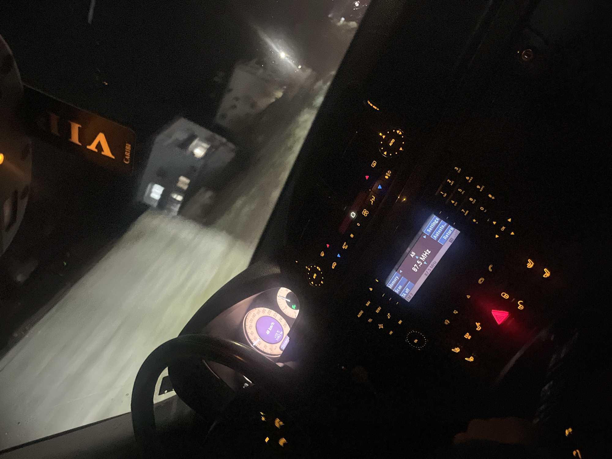 The image is taken from inside a car at night, driving on a road in the city of Srednogortsi, Bulgaria. The driver's side of the vehicle is visible, including the steering wheel, dashboard, and center console, all illuminated. The radio is tuned to 87.5 MHz. Through the windshield, a building is visible on the side of the road, illuminated by the car's headlights. A VIP Caribi card is visible in the front. The speedometer reads approximately 48 km/h. The exterior road is slick, possibly due to rain or snow. The hazard lights are on.