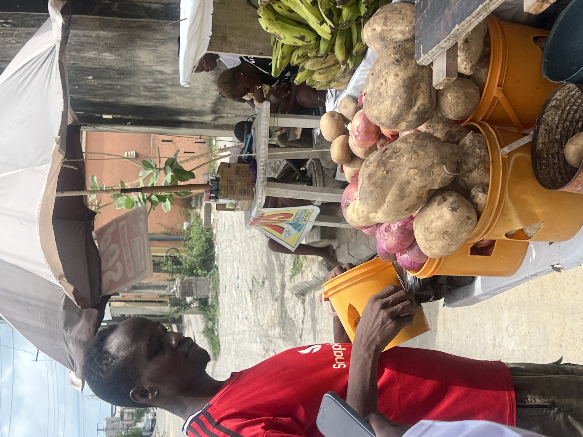 The image captures a vibrant outdoor market or roadside stall scene in Igboefon, Nigeria, during bright daylight.
In the foreground, a dark-skinned man wearing a red short-sleeved t-shirt with a white logo, possibly "Snap!" or "Snappy," and Adidas stripes on the sleeves, is visible. He holds a small yellow plastic bucket in his right hand and a mobile phone in his left, looking towards the market stall.
The market stall, occupying a significant portion of the right side, is laden with fresh produce. Large bunches of green bananas hang prominently above. Numerous large, light brown yams or similar root vegetables are piled high in yellow plastic buckets and arranged on a flat surface, with red onions also visible among them. A white umbrella or awning extends into the frame from the left, providing shade.
Behind the produce, a person with short, light-colored hair is seated, partially obscured by the bananas. This individual, wearing a dark top, appears to be eating or managing the stall. Further in the background, another person is seated on a white plastic stool, with their bare feet visible, seemingly reading a newspaper or flyer placed on the ground, which shows partially legible text like "200" or "2B2".
The background reveals typical urban infrastructure, including buildings with red-orange walls and a concrete ground surface. Green foliage dots the middle ground. The scene portrays a bustling, everyday atmosphere characteristic of a local market, with individuals engaged in daily activities under bright natural light.