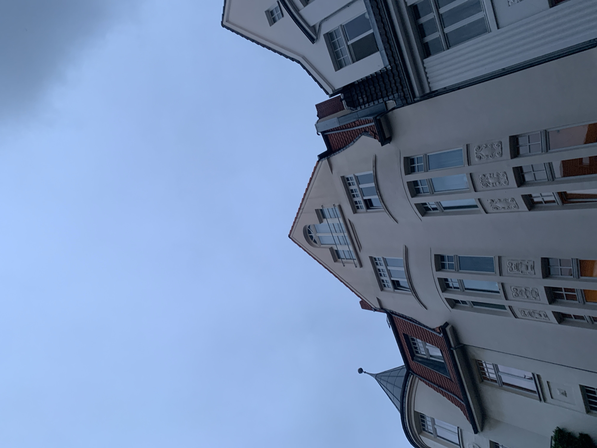 The image presents an architectural view, looking upwards at the upper sections of several buildings against an overcast sky. The photograph is taken at a skewed angle, causing the buildings to appear rotated significantly, almost horizontally across the frame.
The primary subjects are the buildings themselves, which display a blend of classic and possibly Art Nouveau or early 20th-century European architectural styles. They feature light-colored, likely off-white or cream-colored, facades. Notable elements include prominent gabled roofs with reddish-brown tiles and dark eaves. Many windows are visible, including rectangular panes, some with multiple divisions, and a distinctive arched window on one of the central building's gables. One building features a gently curved facade. Decorative elements, such as ornate carved panels between windows on one wall, add intricate detail to the architecture. Another building features a small, conical turret with a dark, pointed roof.
The sky, visible in the background, is a uniform light blue-gray, indicating an overcast or cloudy day. There are no people visible, and no specific activity or event is taking place, making it a static architectural study. The lighting is soft and diffused, consistent with a cloudy daytime setting, possibly morning or late afternoon. There is no visible text. The overall impression is one of solid, well-maintained urban architecture under a muted sky.