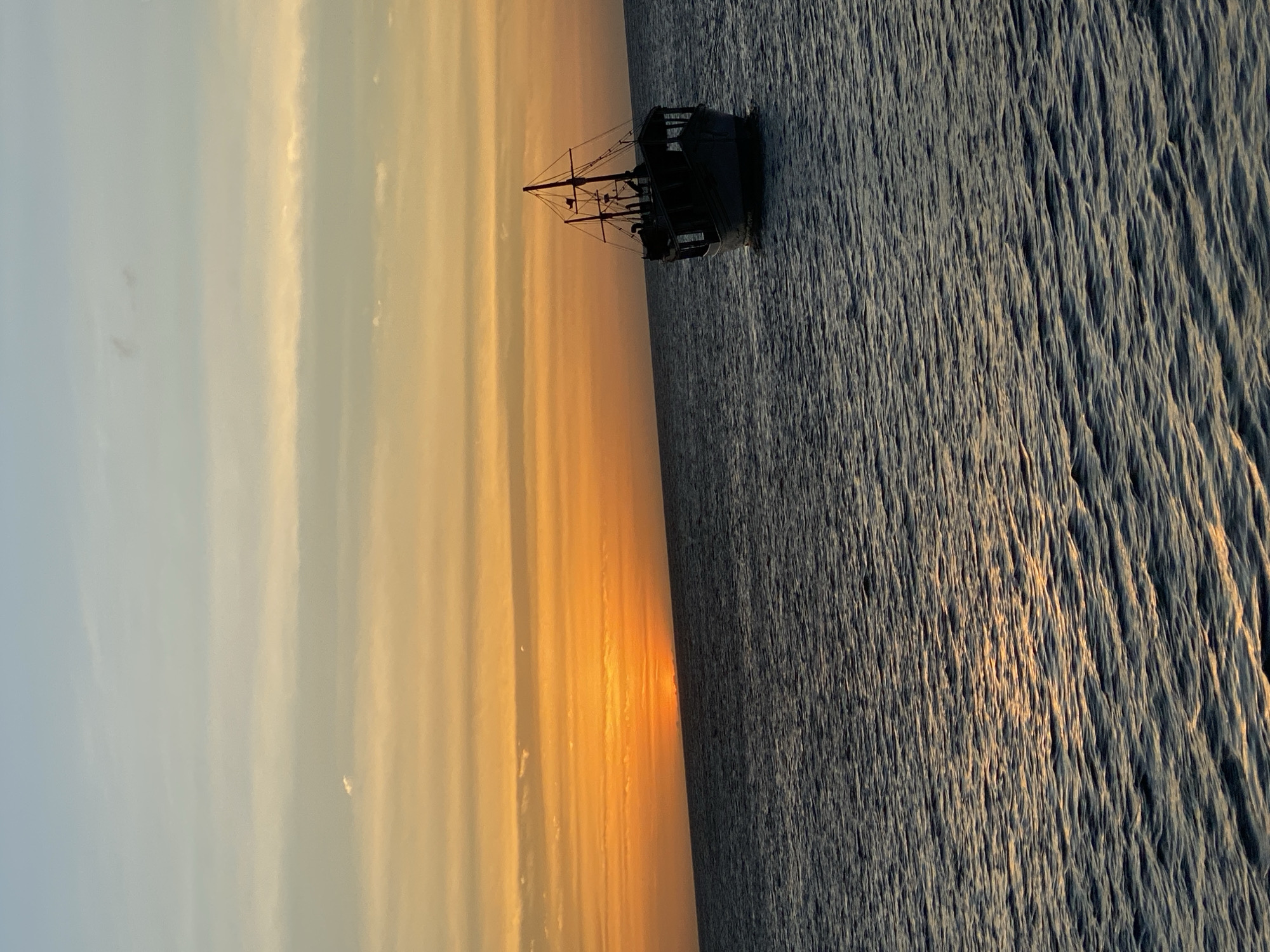The image depicts a sailing ship silhouetted against a sunset at sea. The sky transitions from light blue on the left to orange and yellow towards the center, indicating the setting sun. The sea occupies the right half of the image, its surface textured with waves reflecting the sunlight. The ship, positioned in the center-right, is a dark silhouette with visible masts and rigging. Although the location is indicated to be Juan Griego, Venezuela, there is no specific context that this image reflects its location.
