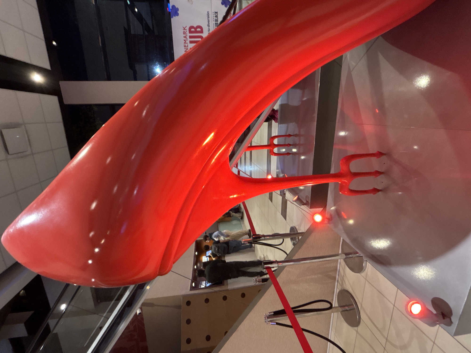 The media file captures a distinctive indoor scene, prominently featuring a large, glossy bright red sculpture. This oversized, stylized object, potentially an abstract high-heeled shoe or a flowing architectural element, dominates the foreground, tilted to the side. Its reflective surface captures ambient light, creating bright highlights.
Beneath the main sculpture, mounted on a white platform, are two smaller red objects resembling devil's pitchforks or tridents, adding a thematic element to the display. Small red lights are embedded into the platform, further accentuating the exhibit. Silver stanchions connected by red ropes cordon off the area, indicating a designated display space.
In the blurred background, several individuals, appearing to be adults, are visible, either observing the display or moving through the space. The setting is a modern, well-lit interior, likely a public commercial building, characterized by tiled floors, glass partitions, and reflective surfaces.
A partially visible banner in the background offers a crucial clue, displaying text such as "...NEMARK UB" and "...tu diversión UB BLACK". This suggests the location is associated with "Cinemark Club" or a similar cinema-related brand, with "tu diversión" (Spanish for "your fun") confirming the context of Buenos Aires, Argentina. The overall impression is that of a contemporary art installation or promotional display within a cinema lobby or entertainment complex. The time of day is not discernible due to indoor lighting.