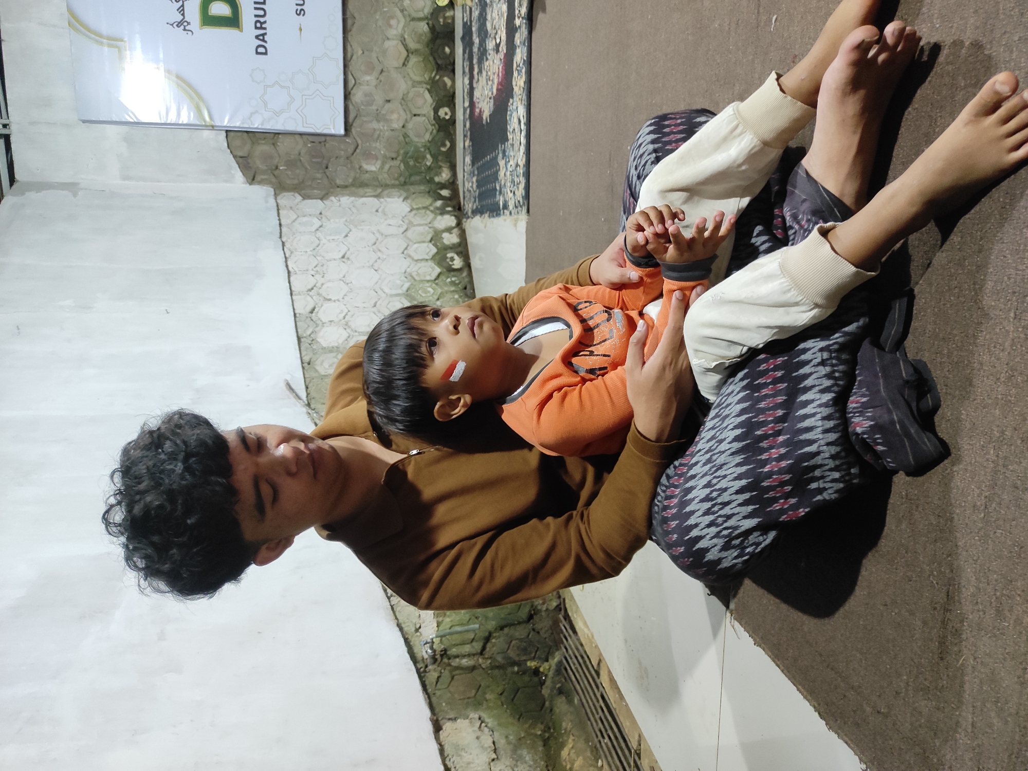 A man and a young child are seated on the floor in what appears to be an indoor gathering space in Sumedang, Indonesia. The man, with curly dark hair, wears a brown zippered shirt and patterned pants. He holds the child on his lap, his arms around the child's torso. The child, wearing an orange long-sleeved shirt and light-colored pants, has face paint on their left cheek, resembling the colors of the Indonesian flag. The background shows a textured wall, a tiled floor, and a mat on the floor. A banner with Arabic calligraphy and text in Indonesian, possibly indicating an event or location, is visible on the wall. The lighting suggests an evening setting.