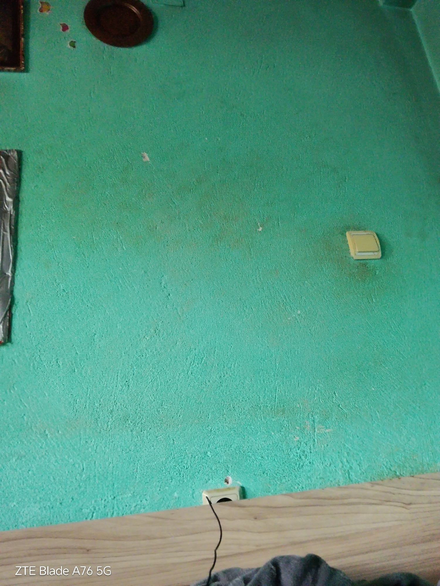 The scene, presented as being located in Stara Zagora, Bulgaria, depicts an indoor wall with a rough, textured finish, painted in a faded teal or greenish-blue hue. Visible discoloration, appearing as brownish-grey stains or dirt, is present across the wall surface, particularly around the center and near the electrical fixtures.
In the upper left quadrant, a portion of a framed object, possibly a picture or mirror, is visible, featuring a dark border. Above the center-top, a dark brown, circular, shallow object, resembling a decorative plate or bowl, is mounted directly on the wall. Below this object, three small, colorful, heart-shaped decorative elements are affixed to the wall.
Towards the right-center, a light yellow or beige wall-mounted electrical switch or cover is present, with minor brown discoloration immediately beneath and around its base. In the lower-left section of the wall, a white electrical outlet is visible, from which a black cable extends towards the right. This cable rests upon a light-colored wooden surface, which occupies the lower portion of the image, displaying a visible wood grain pattern. Partially visible in the lower right, resting on the wooden surface, is a dark grey, textured fabric item.
The timestamp "ZTE Blade A76 5G" is superimposed in white text on the lower left of the image. No individuals or actions beyond the presence of stationary objects are depicted.
