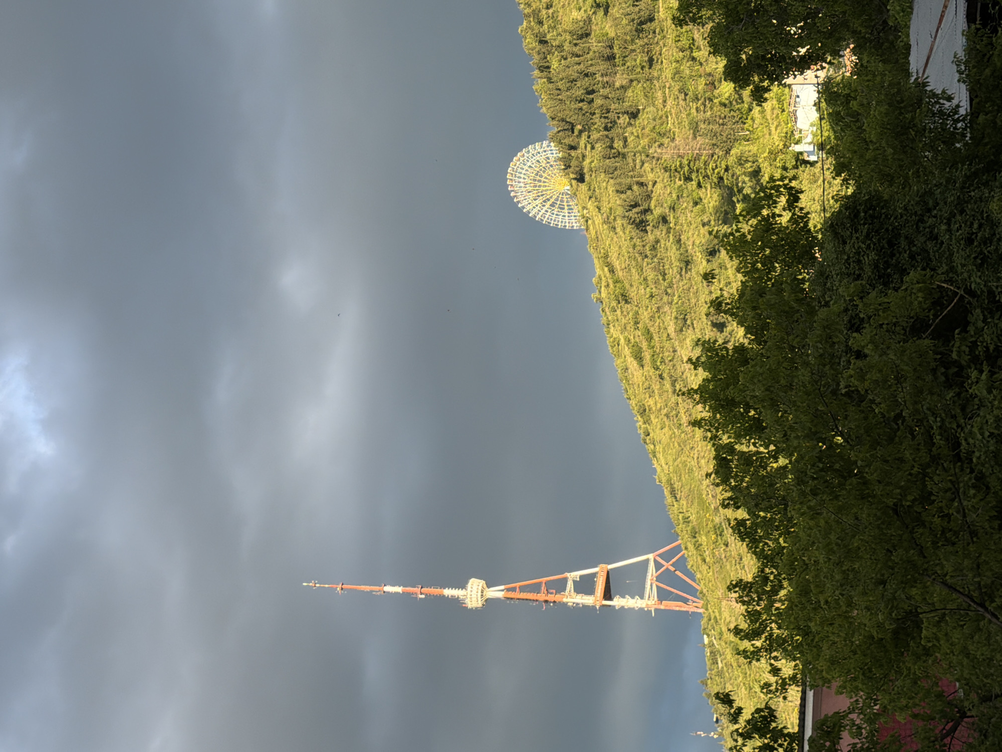 The image depicts a view in Tbilisi, Georgia. In the foreground is the Tbilisi TV Broadcasting Tower, a tall, red and white structure extending upwards into a stormy, overcast sky. Adjacent to the tower and slightly behind it is a heavily wooded hillside. At the top of the hillside, a large Ferris wheel is visible. Some buildings are partially obscured by trees at the top of the frame. Green trees and foliage frame the right side of the image.