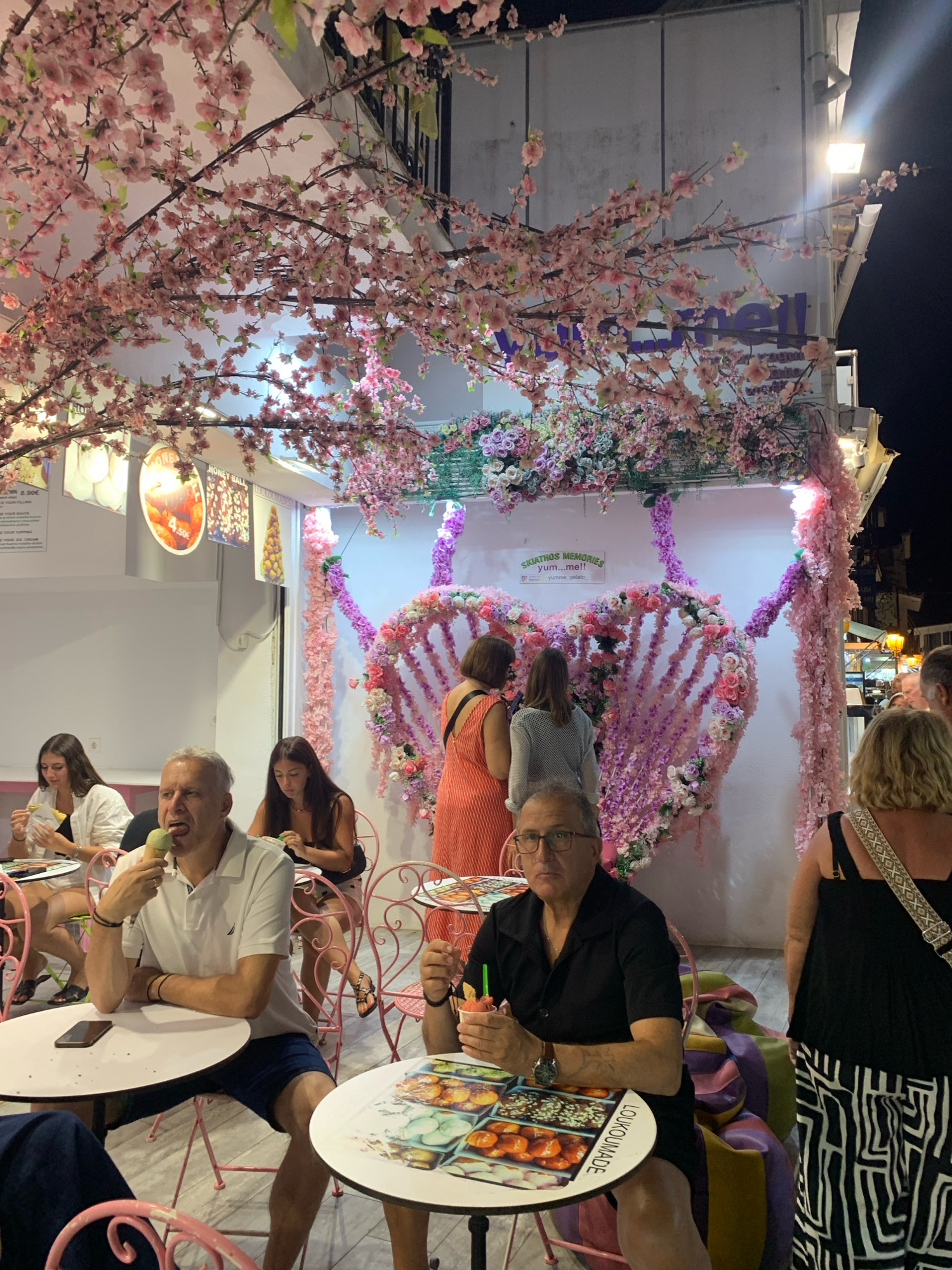 This is a medium shot, taken at eye level, of an outdoor seating area of a gelato shop decorated with artificial cherry blossoms and floral arrangements, located in Skiathos, Greece. In the foreground, a middle-aged man wearing glasses, a black short-sleeved collared shirt, and dark shorts sits at a round white table with a pink metal chair. He is holding a small cup with a green straw in his right hand and looking towards the camera. Another middle-aged man, also wearing glasses, sits at a similar table with a pink metal chair to his right and is eating green ice cream from a cone, looking to his left. In the background, several women are seated at tables. One woman in an orange dress is standing, and another woman in a white top and dark shorts is seated. The shop's facade is decorated with a large heart-shaped floral arrangement composed of pink and purple flowers, and a sign reads "SKIATHOS MEMORIES yum...me!!". Overhead, branches of artificial pink cherry blossoms extend across the scene, with lights from the shop illuminating the area. The overall atmosphere is festive and illuminated for evening patrons.