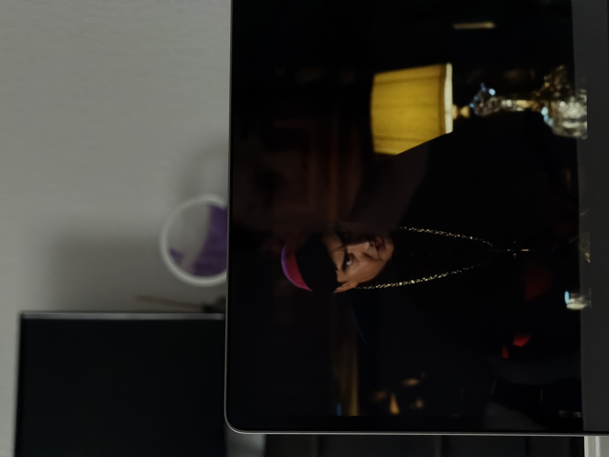A man wearing a black cassock and a red zucchetto is visible on a laptop screen, looking intensely to his right. He has dark hair and is wearing a gold chain with a cross. To the right of the man on the screen, a lamp with a yellow shade sits on a table. The laptop is placed on a surface in front of a blurry background that includes a dark rectangular object on the left and a wall with a circular object on the right. The scene appears to be indoors. The location is not specified.