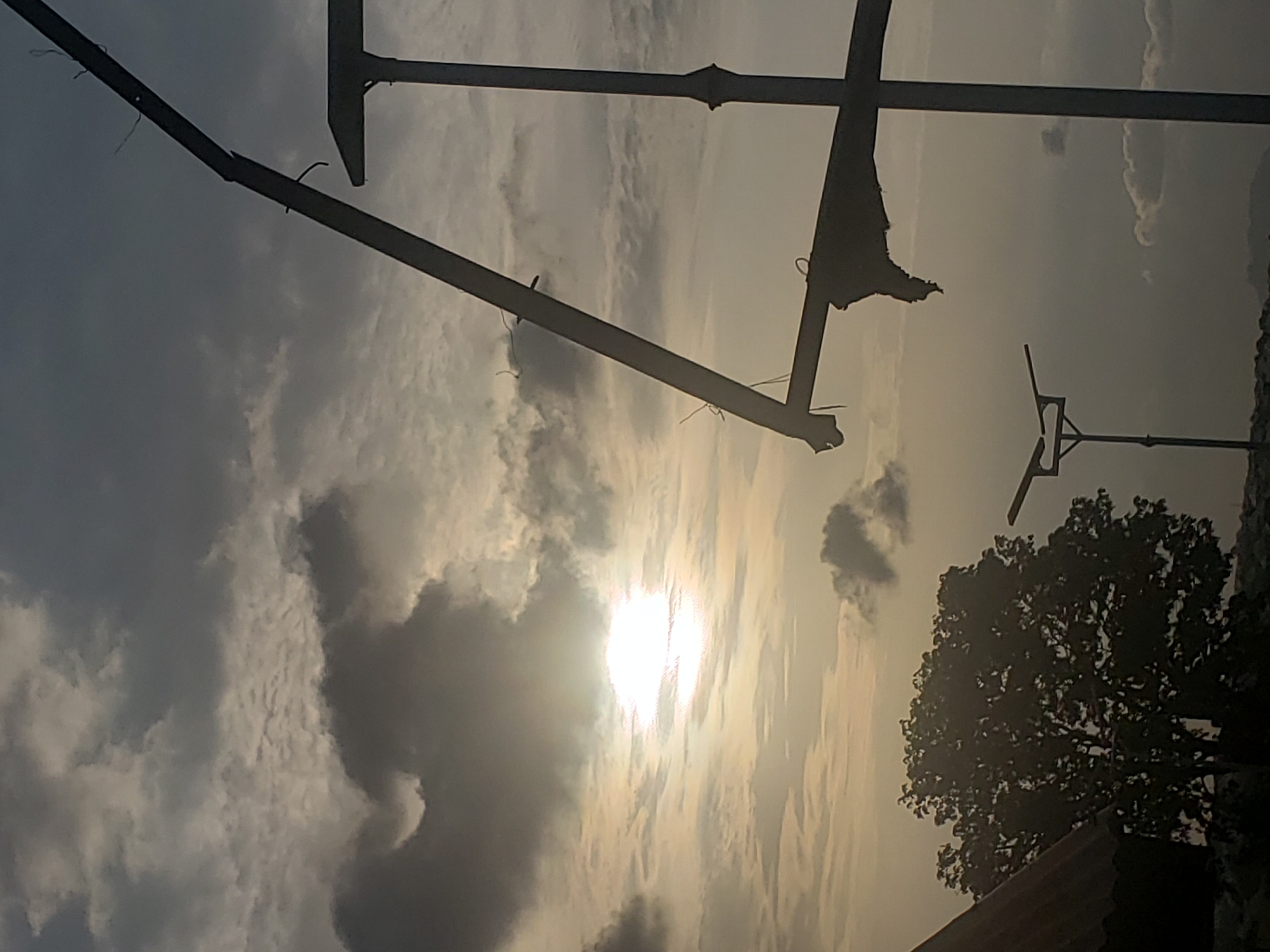 A landscape at dusk in Adankolo, Nigeria, featuring a silhouette of trees on the right side and a bright sun obscured by clouds in the center. Overhead, a metallic structure spans across the top, appearing to be construction or utility infrastructure. A second antenna-like structure is visible to the right of the tree. The overall color palette is muted with shades of gray and gold.