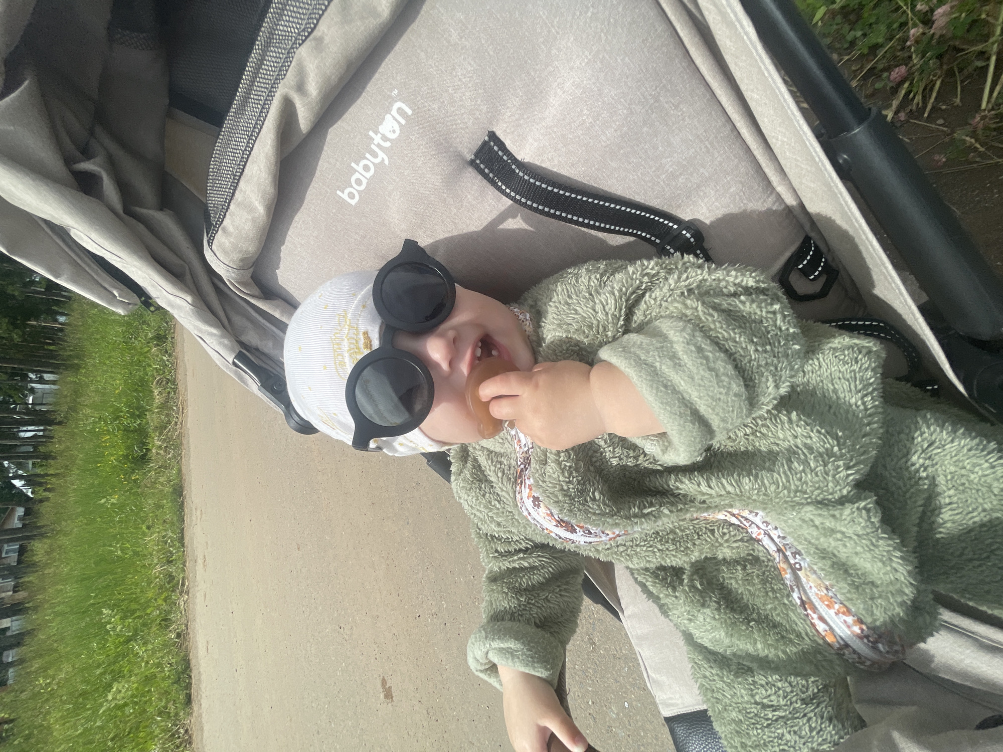 A baby is lying in a stroller, wearing a green fuzzy onesie and a white cap with a gold pattern. The baby is also wearing large, round black sunglasses and has a pacifier in its mouth. The stroller has a light brown fabric with the brand "babyton" visible in white text. Straps with black and white stitching are attached to the stroller. The background shows a paved path next to a grassy area with trees and buildings, suggesting an outdoor setting during the daytime with sunny weather. The baby appears content.