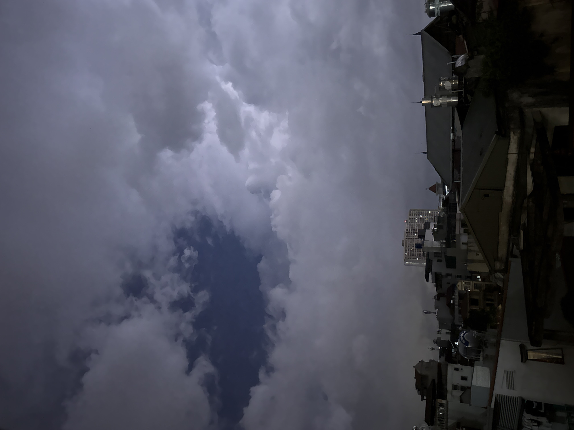 The image depicts a nighttime urban scene, likely in Hà Nội, Vietnam, given the architectural style of the buildings. The sky is dominated by heavy, dark clouds, with a small patch of darker blue sky visible. The urban landscape consists of densely packed buildings, a mix of low-rise and a few taller structures. Rooftops are cluttered with water tanks and other utilities. The image is rotated counter-clockwise.