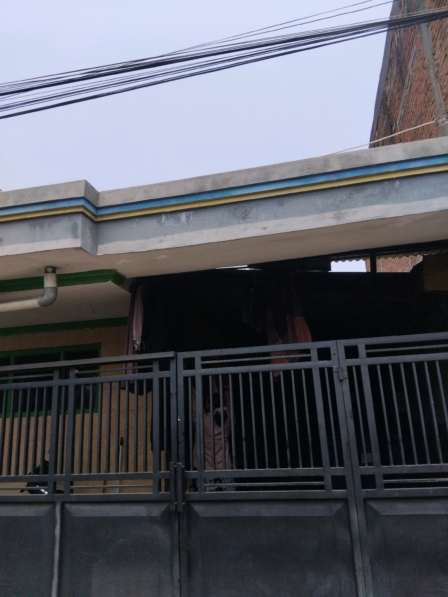 The image shows a view of a property's exterior. In the foreground, a dark gray metal gate with vertical bars is visible. Behind the gate, a black metal fence with square openings is also present. Through the fence and gate, a portion of a building is partially visible, featuring green-framed windows. The roofline of the building is adorned with a gray, yellow, and blue decorative trim. A white PVC pipe extends from the underside of the roof. The sky above is overcast and appears light gray. Overhead, a cluster of black electrical wires runs horizontally across the frame. To the right, the corner of a red brick building is visible, extending upwards. The scene is likely a residential or commercial property in Kepuh Kiriman, Indonesia, given the architectural style and the presence of electrical wires.