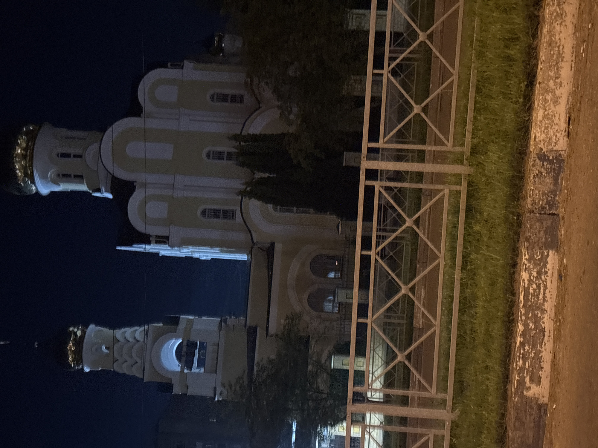 The image shows a dimly lit night scene of the Holy Trinity Cathedral in Obninsk, Russia. The cathedral is illuminated, revealing its white and yellow facade, domes, and a bell tower. A metal fence with a diamond pattern is in the foreground, separating the viewer from a strip of grass and then the street. The sky is dark. There are no people or vehicles visible in the image.