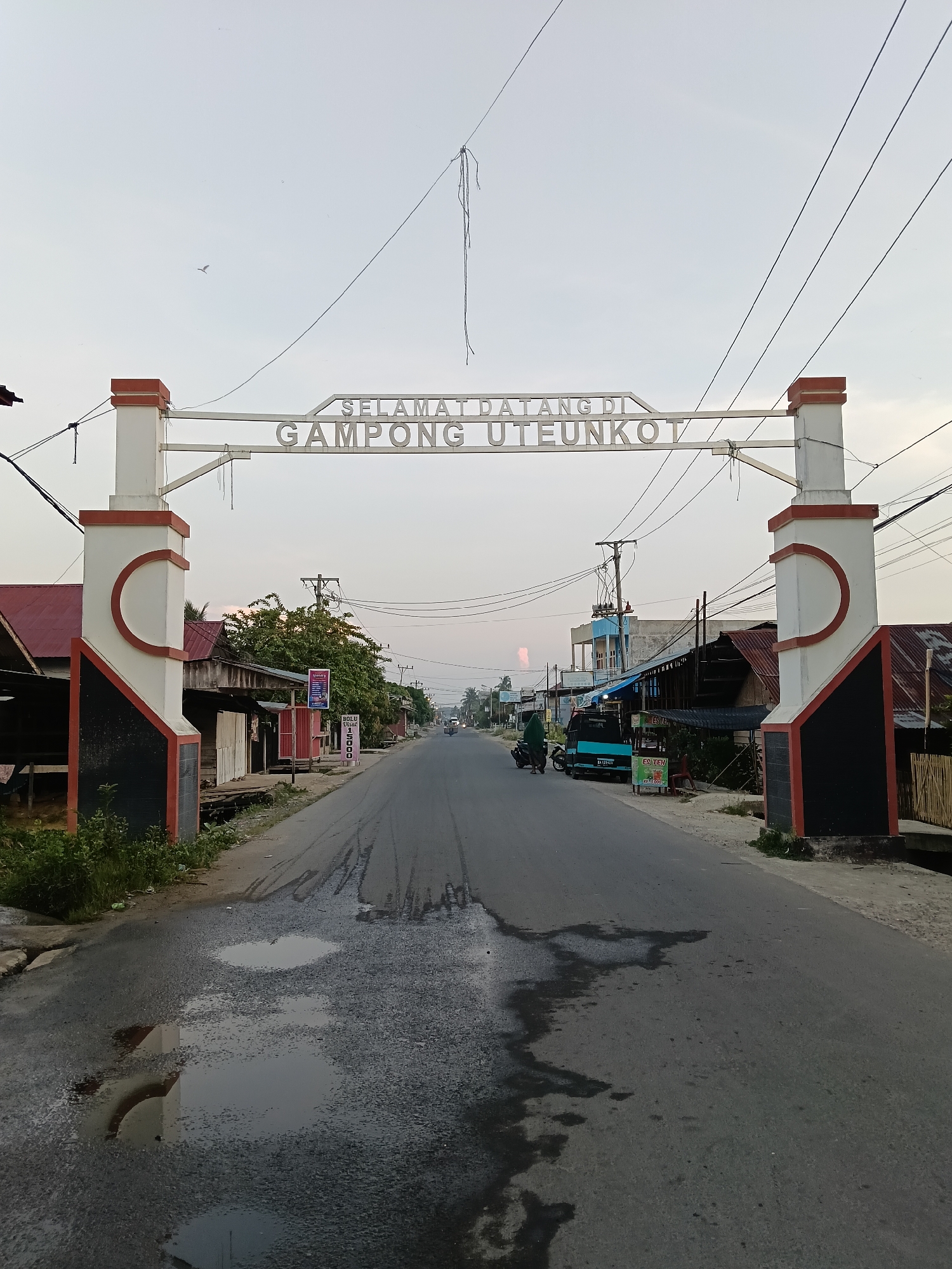 The image depicts the entrance to "Gampong Uteunkot" in Lhokseumawe, Indonesia. A large archway with white pillars and a sign in Indonesian spans the road. The pillars have orange trim at the top and black paint at the base. The road is asphalt with areas of standing water. Buildings line both sides of the street, appearing mostly as single-story structures. Power lines are strung overhead. A blue van is parked on the right side of the road. A person dressed in green stands beside the van. A motor scooter is moving along the street in the distance. The sky is overcast.