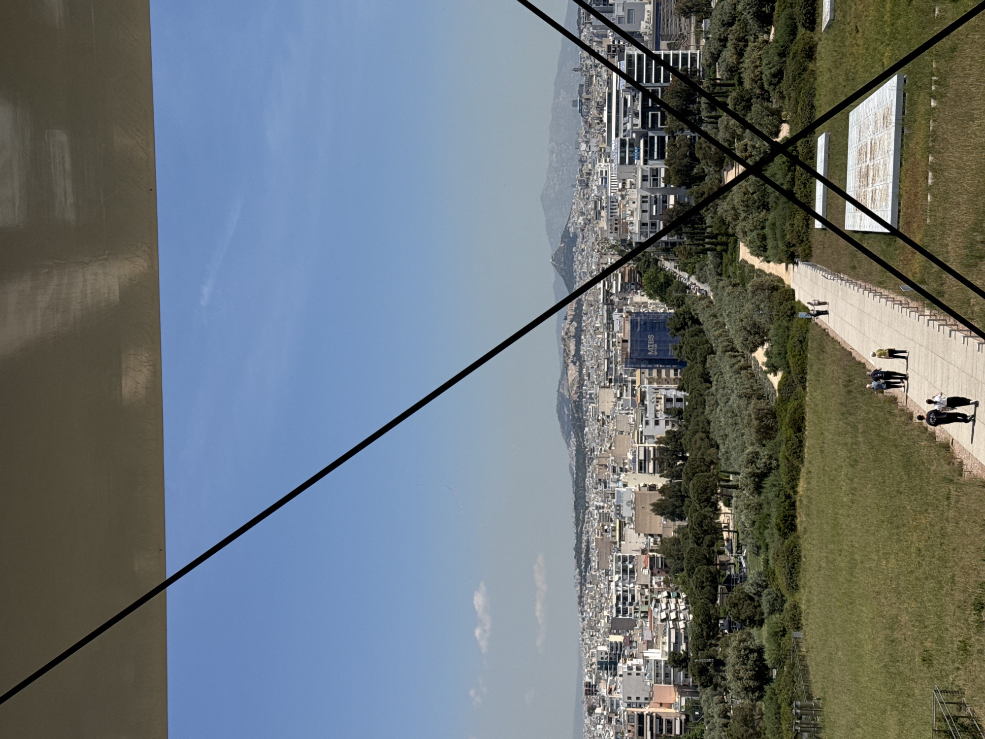 The image captures a panoramic view of Kallithea, Greece, under a clear, bright blue sky on a sunny day. The scene appears to be taken from an elevated vantage point, possibly a building or platform, evidenced by a large, light-colored structure occupying the left portion of the frame and diagonal black lines, likely cables or structural supports, crisscrossing the view.
In the mid-ground, the sprawling urban landscape of Kallithea unfolds, characterized by numerous white and light-colored buildings with distinct Mediterranean architectural styles, extending towards a hazy mountain range in the distance. A large, dark blue building with "MIBS group" text visible on its side stands out amidst the urban density.
The foreground features a wide expanse of vibrant green grass, bordering a paved pathway. Several people are visible strolling along this path, which is lined with small bollards or posts. To the right of the pathway, on the grassy area, are two large, rectangular structures made of white panels or tiles, possibly part of an outdoor installation or solar array. The general atmosphere suggests a peaceful, clear day, offering an expansive view of the city and its natural surroundings.