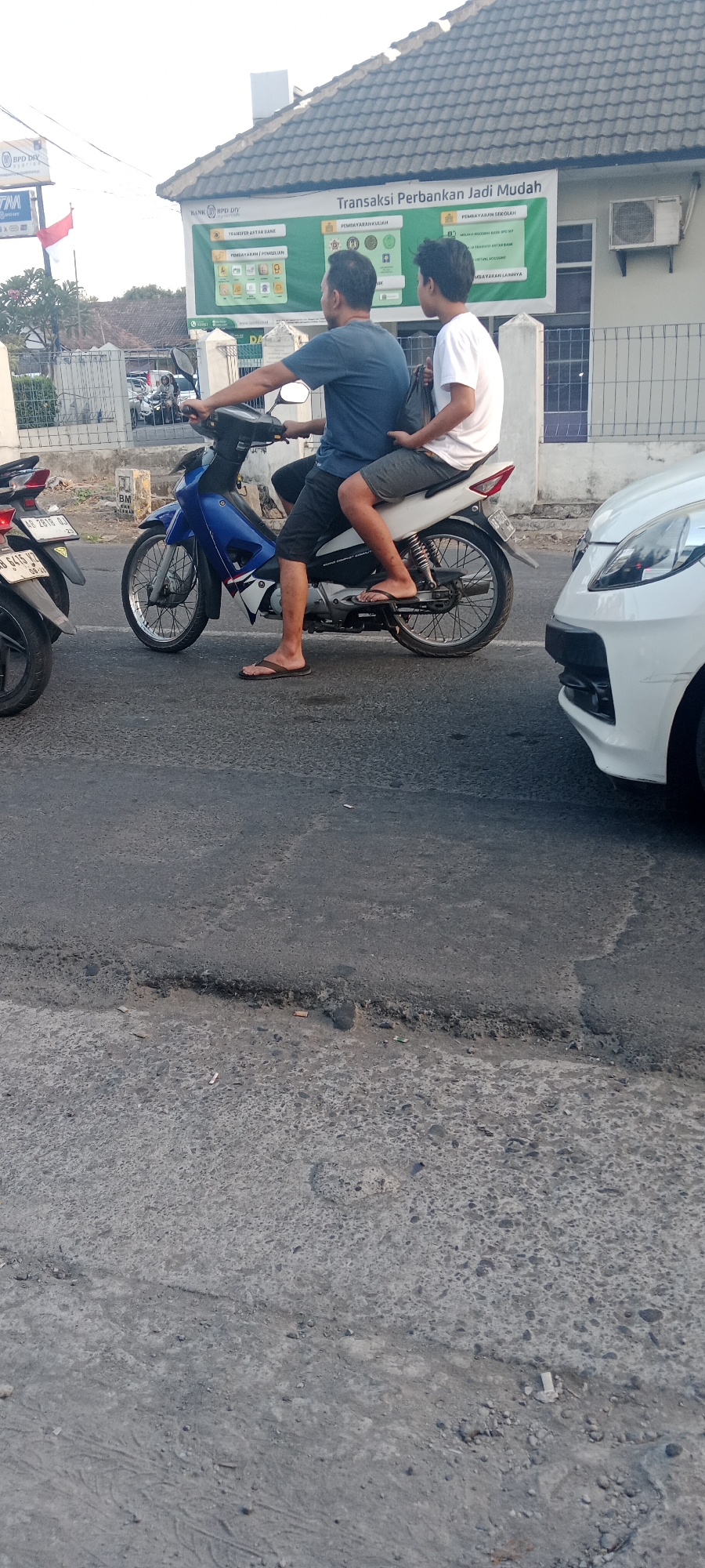 Two individuals are riding on a blue and white motorcycle on an asphalt road in Maguwoharjo, Indonesia. The person in front is driving, wearing a dark blue t-shirt and dark shorts. The person behind is wearing a white t-shirt and grey shorts, holding a dark bag. The motorcycle has a license plate that reads "AB 2818 UJ". A white car is visible to the right of the motorcycle. In the background, a building with a sign that says "Transaksi Perbankan Jadi Mudah" and "BANK BPD DIY" is present, along with a white fence and some trees. The sky is bright, suggesting daytime. The road surface appears worn with some patches of gravel and debris.
