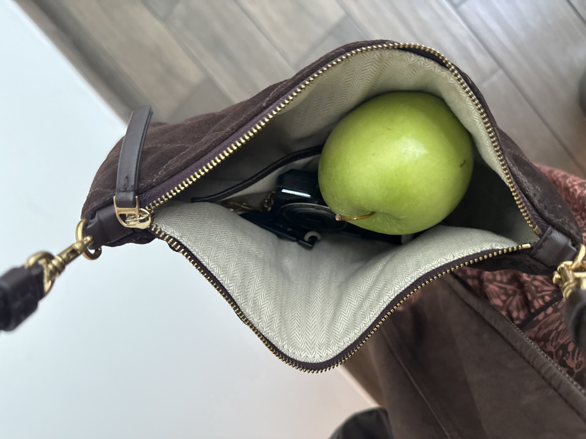 A partially opened brown suede purse with a light-colored patterned lining contains a green apple and what appears to be keys and other small items. The purse's zipper is partially closed. The background shows a combination of light and dark surfaces, possibly a floor and other parts of the purse. Location details are not discernible from the image.