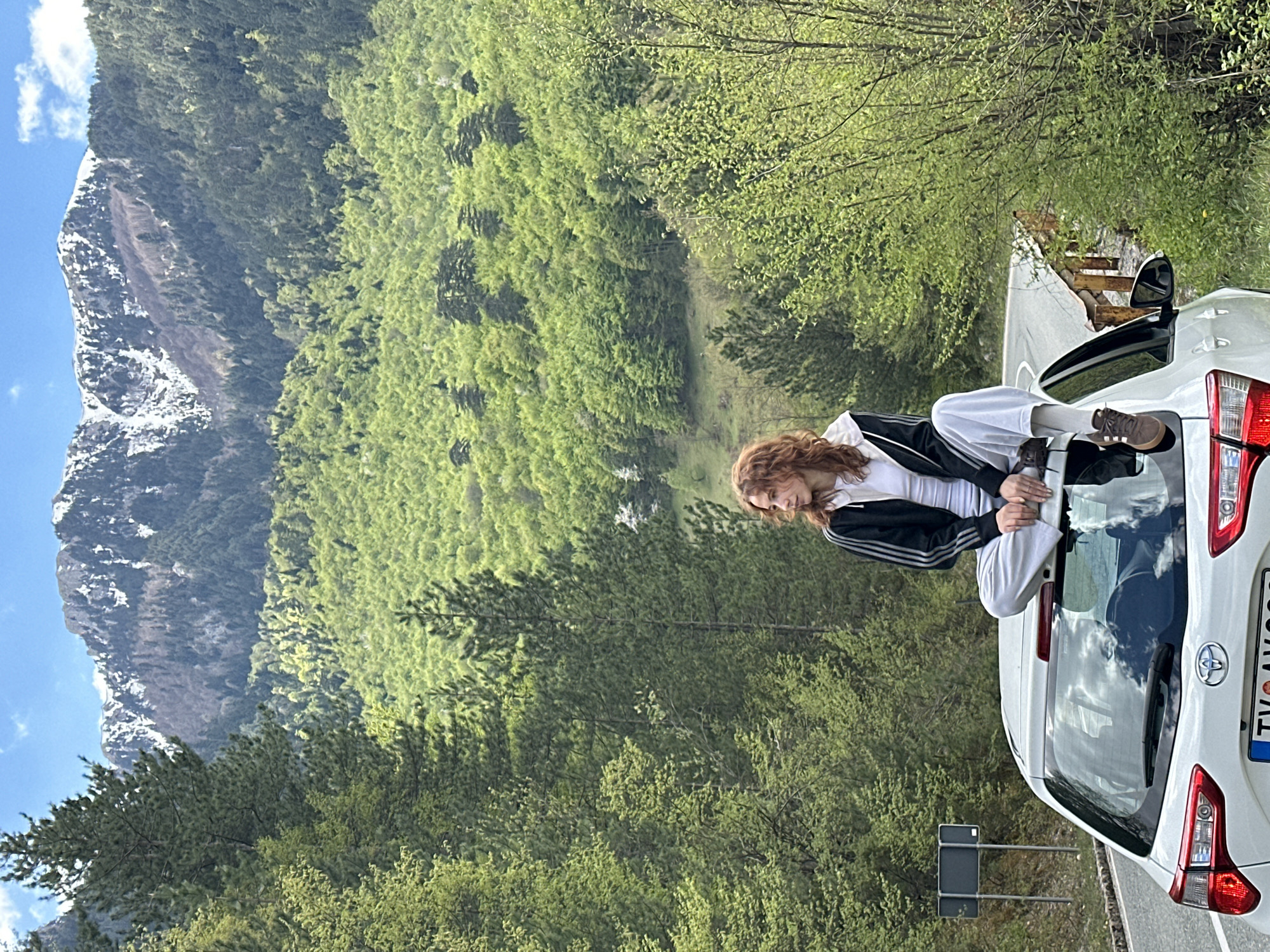 A young woman with red hair is sitting on the open trunk of a white car on a road. She is wearing a white t-shirt, a black Adidas tracksuit jacket, and white pants. Her legs are bent and she is holding onto the edge of the trunk with her hands. The car is parked on a paved road.
In the background is a lush, green mountain covered in trees. There is a taller, snow-capped mountain visible in the distance. The sky is partly cloudy. The license plate of the car is partially visible and appears to be European. There is a road sign behind the car.
The location appears to be Bistrica, Montenegro based on the landscape and license plate style.