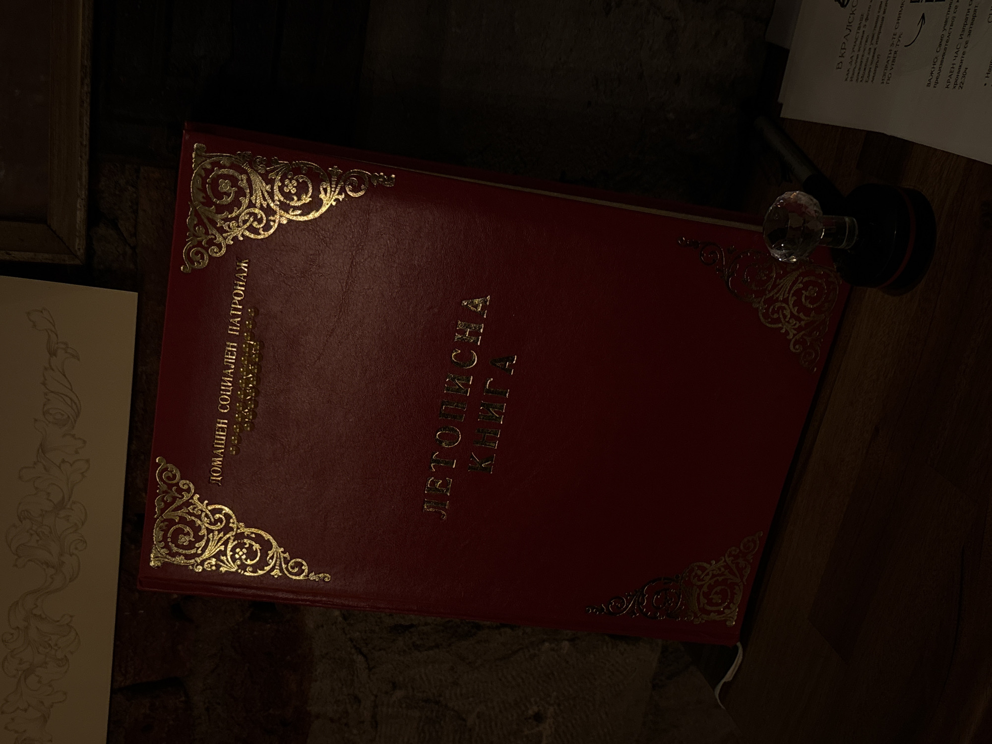 The image shows a red book titled "Літописна книга" (Letopisna kniga), which translates to "Chronicle Book", with the inscription "ДОМАШЕН СОЦИАЛЕН ПАТРОНАЖ" (DOMASHEN SOCIALEN PATRONAZH), meaning "Home Social Patronage", in gold lettering on its cover. The book has decorative gold corner embellishments and rests on a surface that appears to be stone or textured material. To the right of the book is a glass and black ink stamp, and part of a printed page is visible, with text that mentions "В КРАЛСКО" (V KRALSKO) and instructions on how to participate by submitting photos via Viber, along with a deadline. The context suggests the book and its placement are in Bulgaria, possibly Varna, based on the Cyrillic script and content on the page.